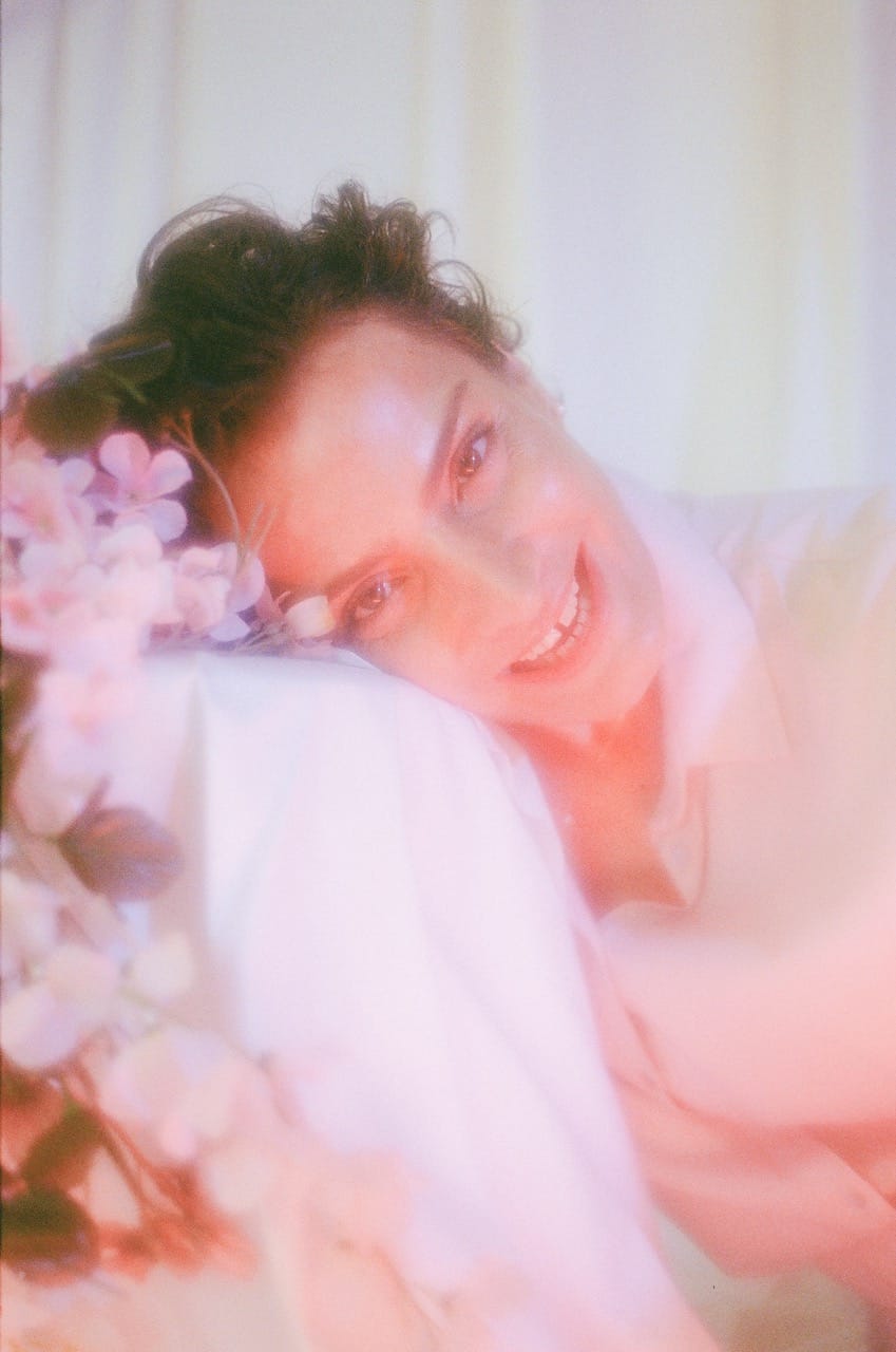 A person smiling, resting their head on a white surface, surrounded by soft pink flowers and a dreamy, pastel background.