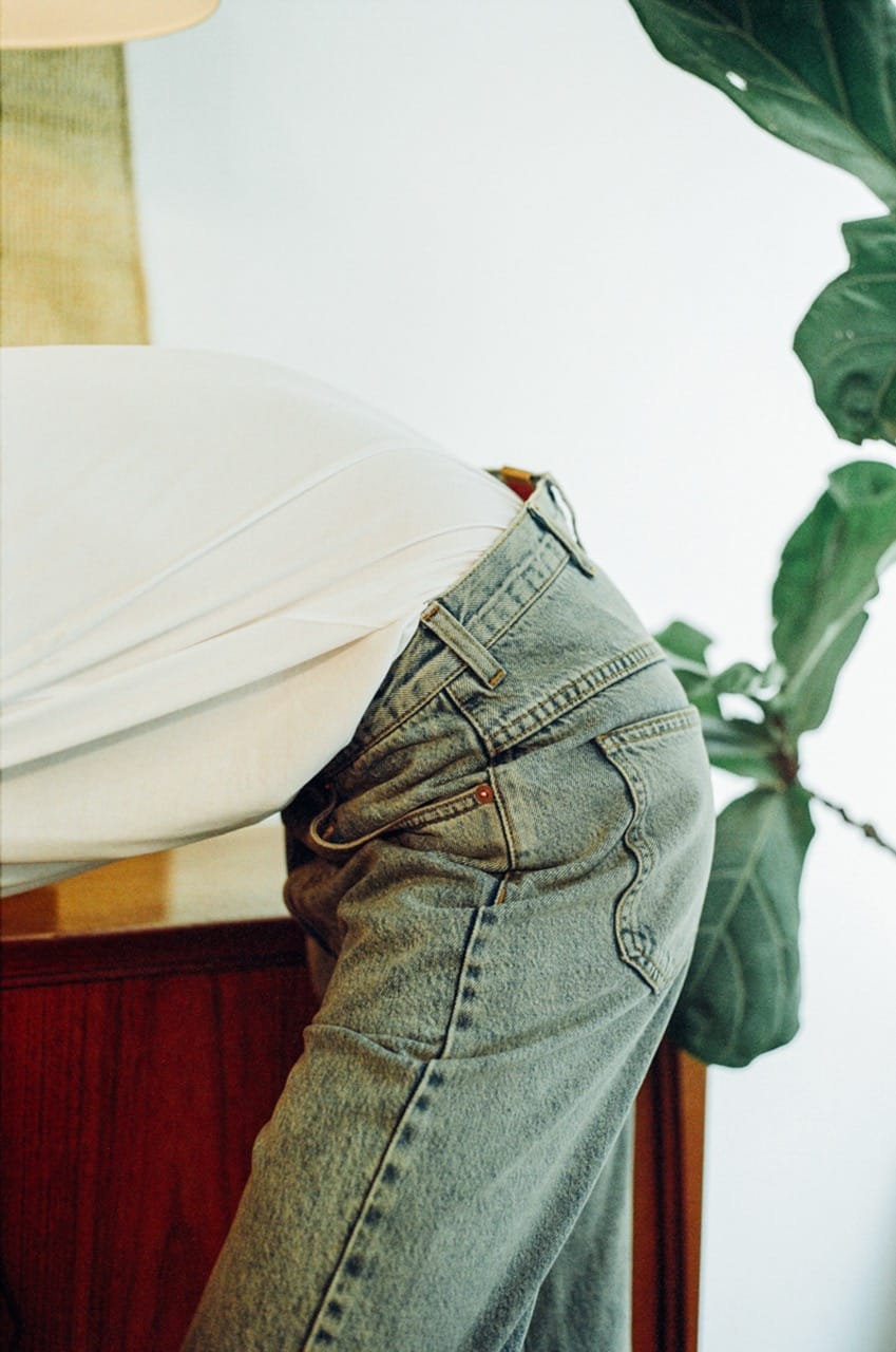 Person in a white shirt and jeans leaning over a wooden surface, next to a large leafy plant.
