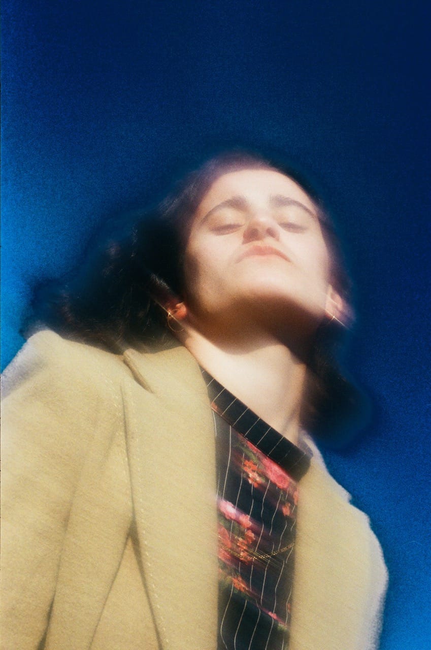 Artistic photo of a person in a beige coat, head tilted back, with a blurred effect against a deep blue background.