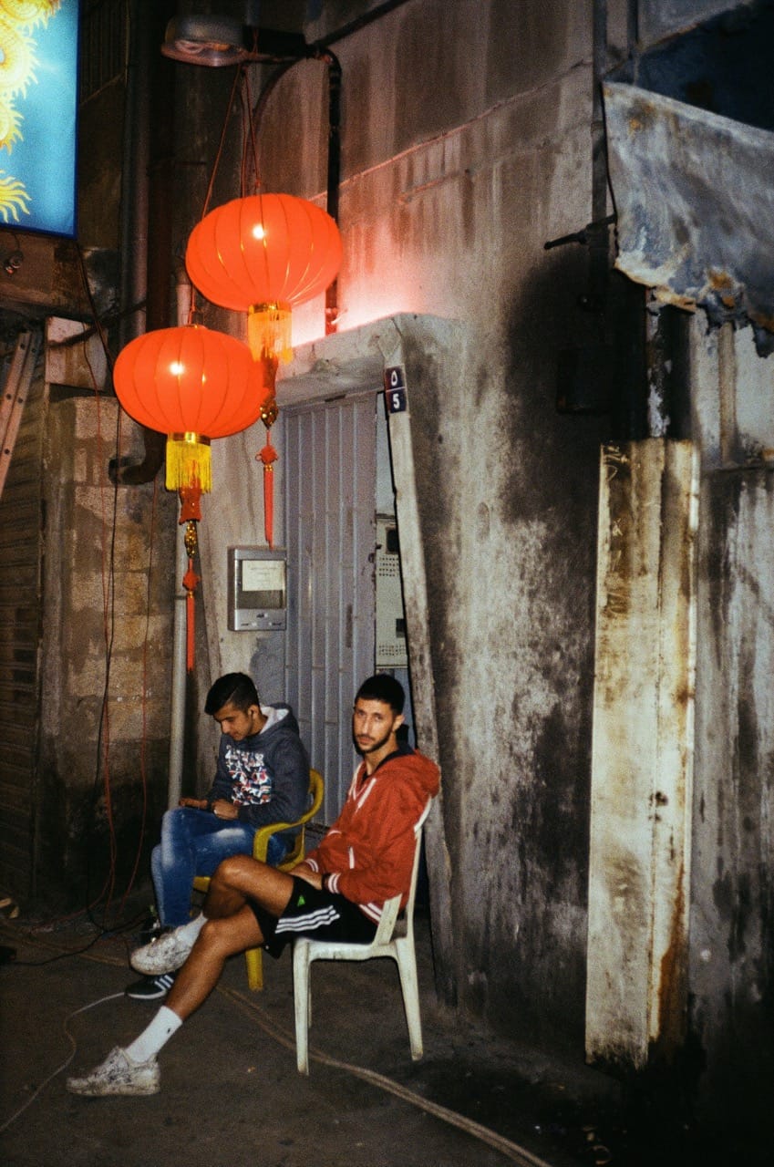 Two men sit on plastic chairs outside a building at night, under red lanterns. One uses a phone, the other looks ahead. Urban setting.
