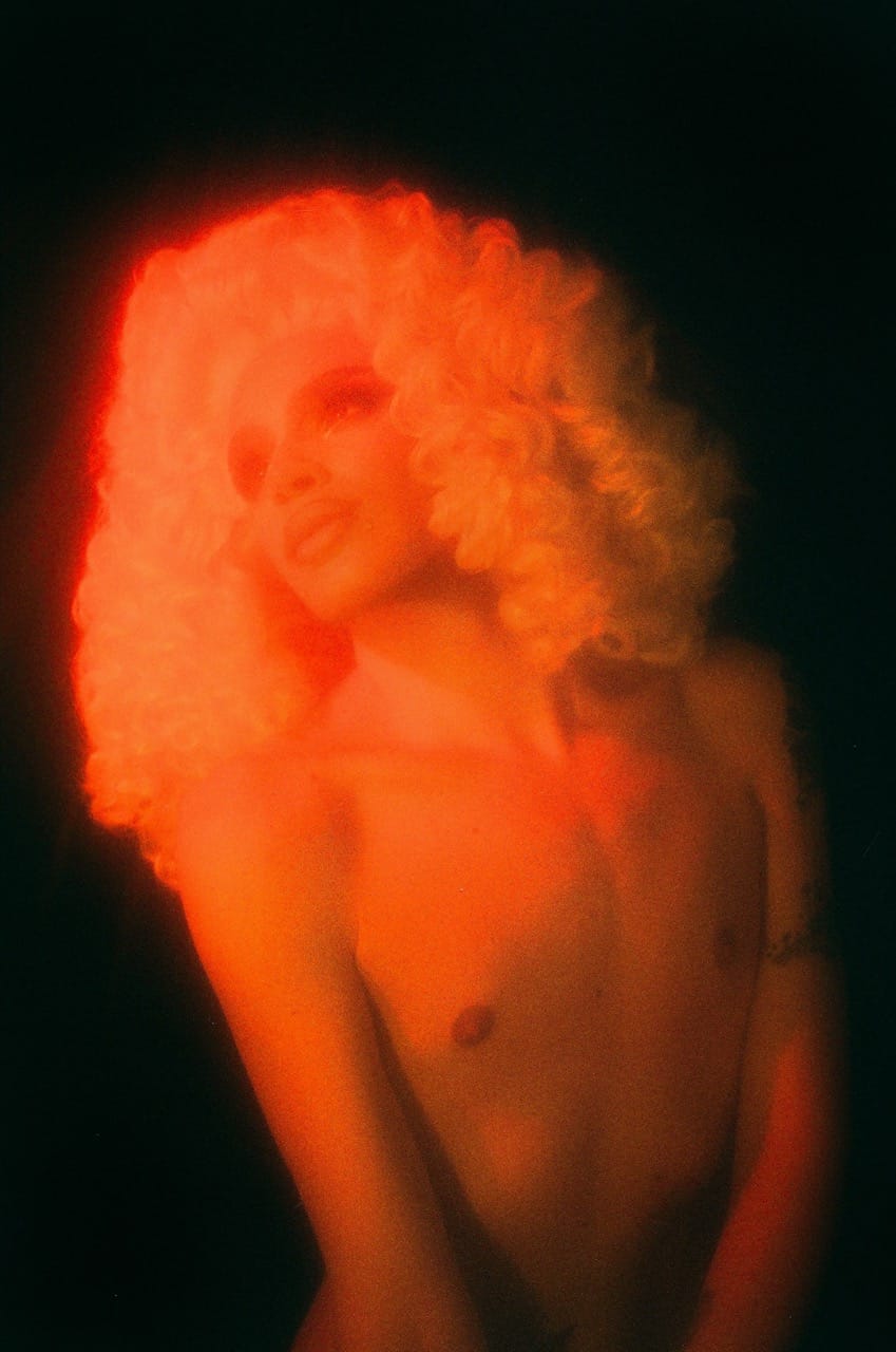 Person with voluminous curly hair, illuminated by red light, posing against a dark background.