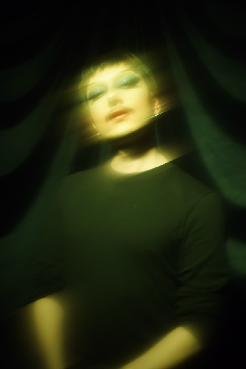 A person with blurred motion effect, wearing blue eyeshadow and a black top, stands against a dark, draped background.