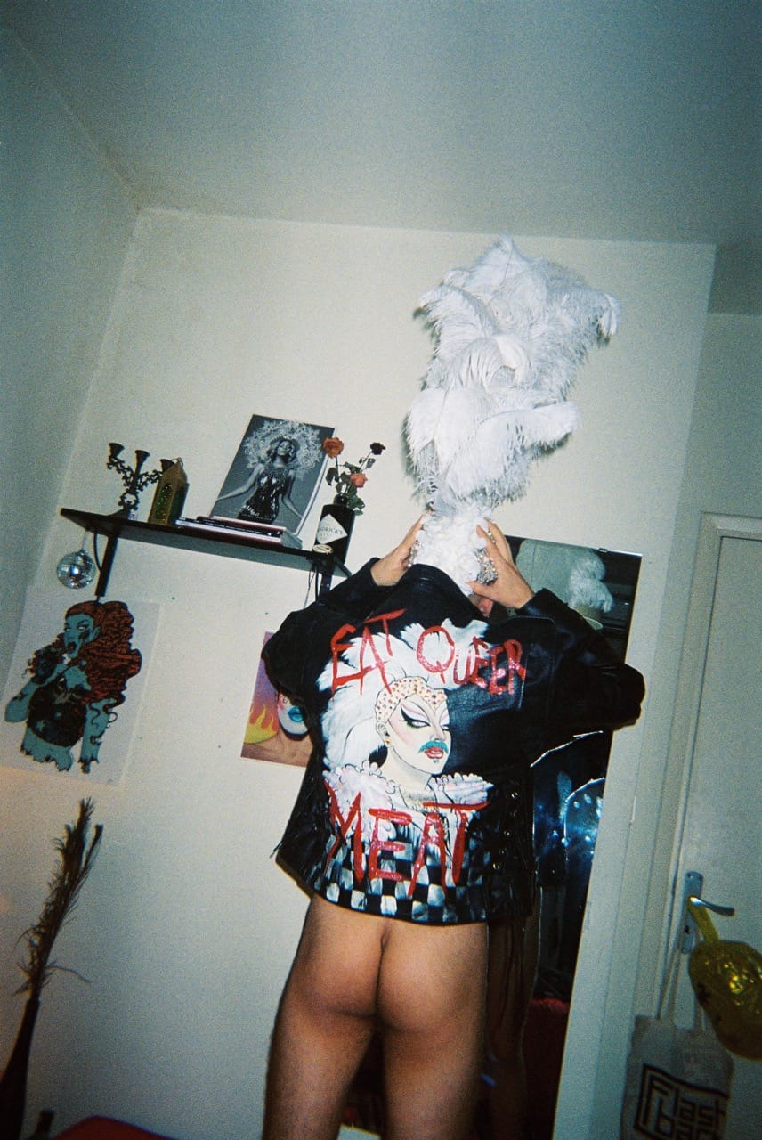 Person wearing a jacket with "Eat Queer Meat" design, holding a feathered headpiece, standing in a room with art on the walls.