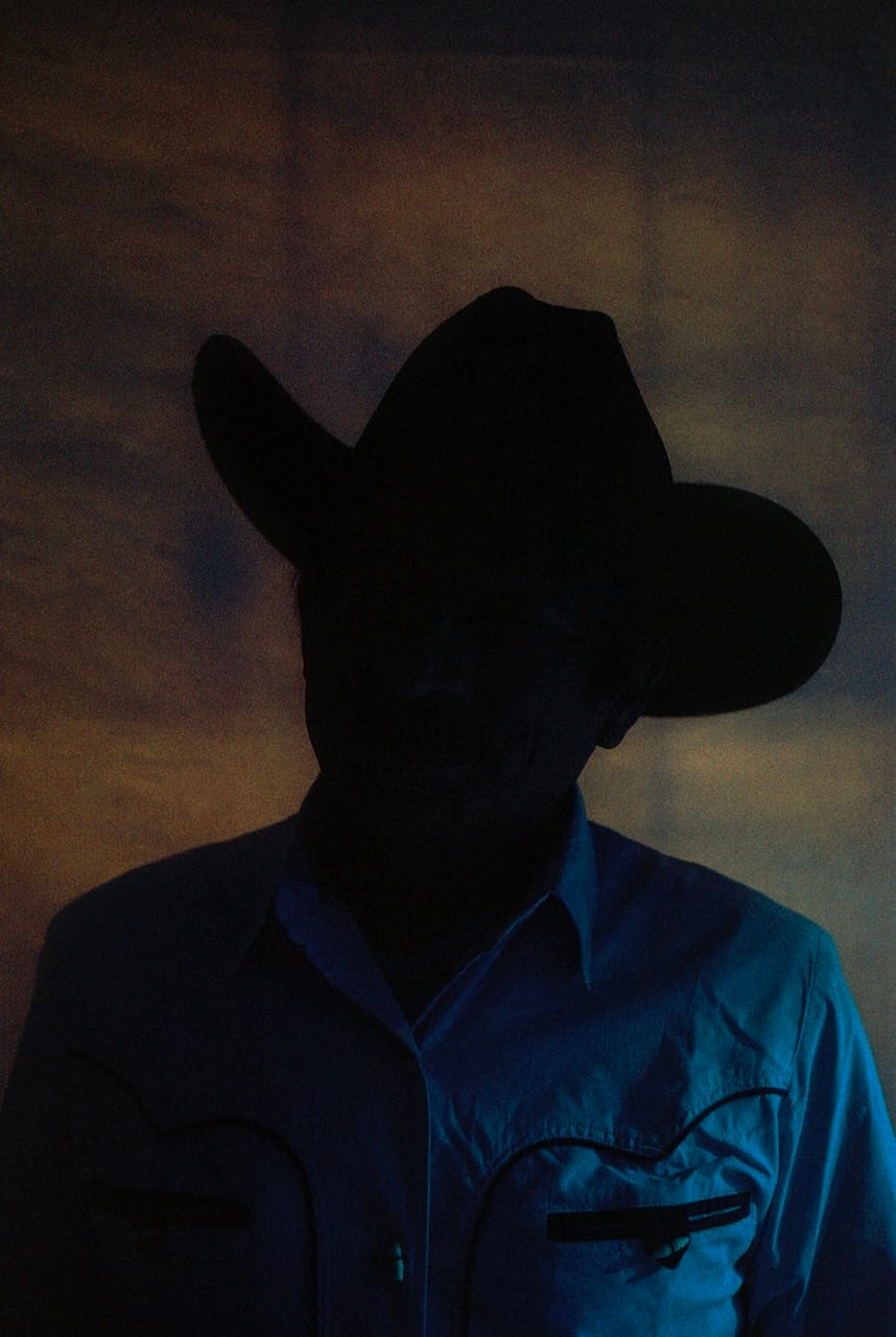 Silhouette of a person in a cowboy hat and western shirt against a softly lit, textured background.