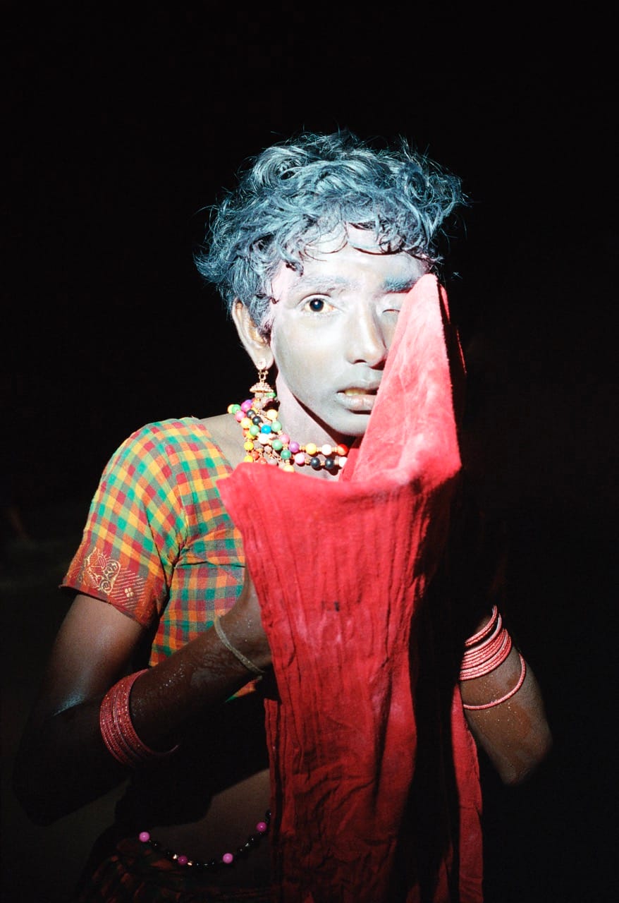 Person with painted face and colorful attire partially covers face with a red cloth, standing against a dark background.