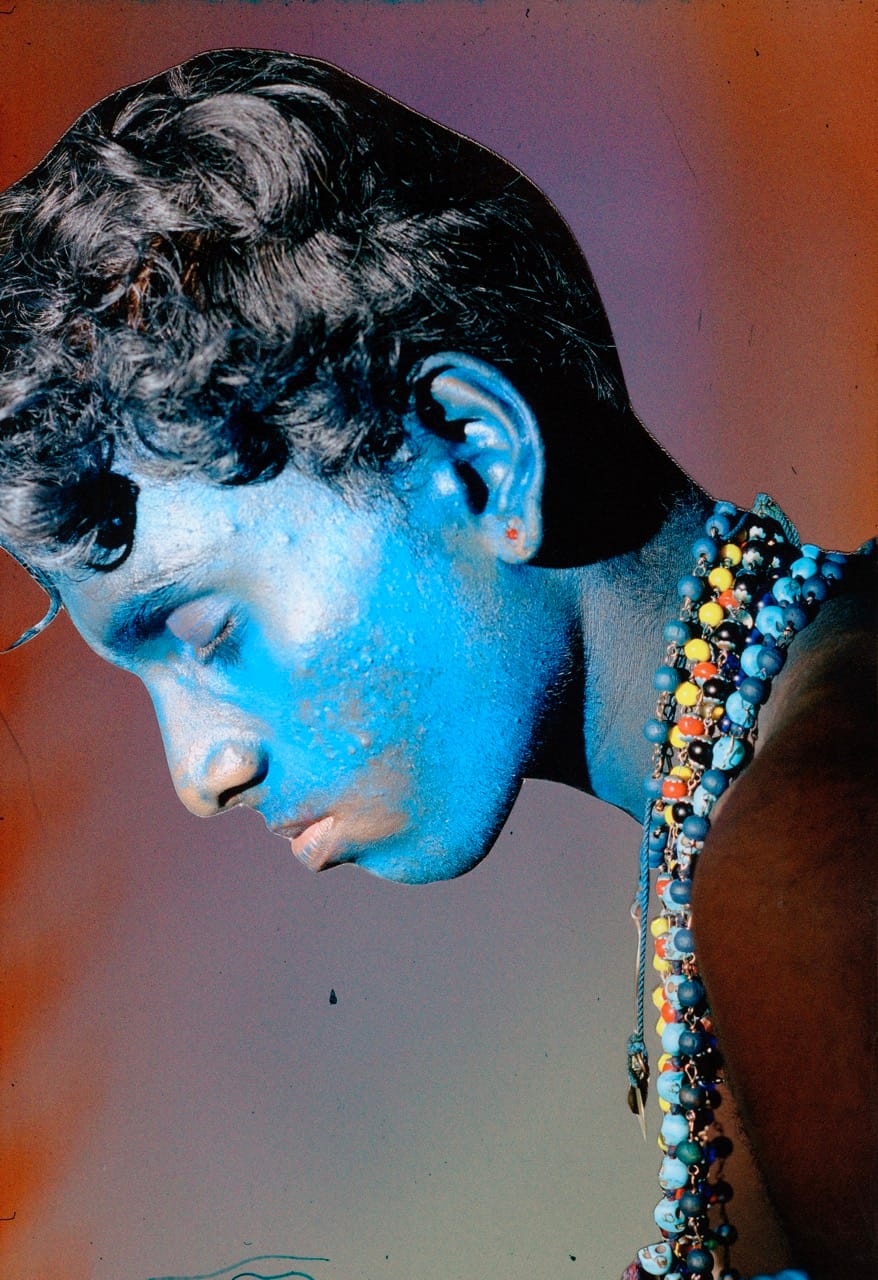 Person with blue-painted face, eyes closed, wearing colorful beaded necklaces against a gradient background.