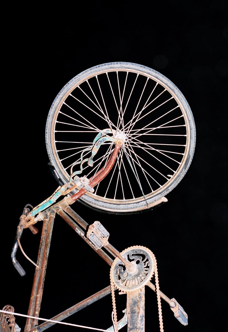 A rusty bicycle with a bent front wheel is suspended against a dark background, highlighting its worn and weathered appearance.
