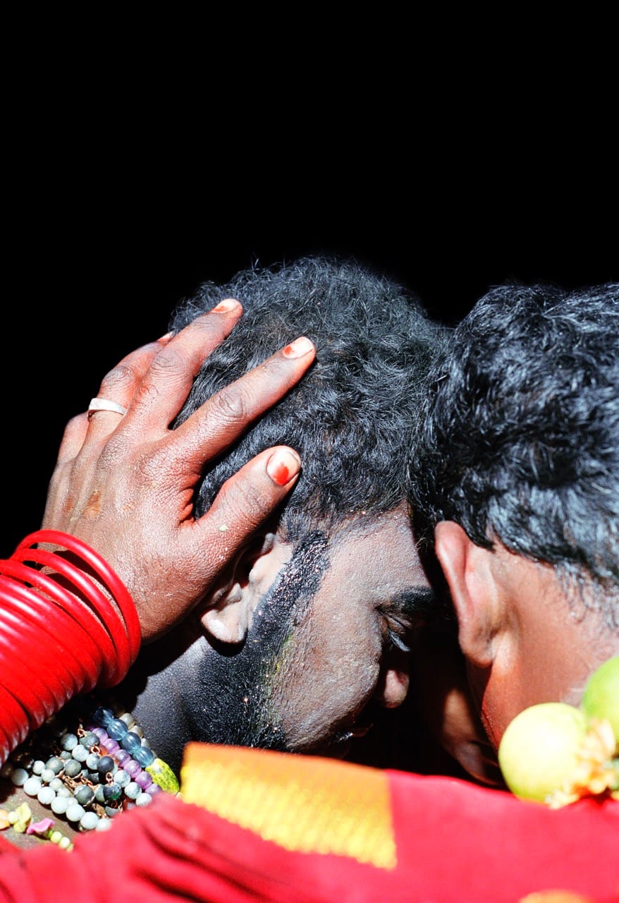 Two people in close embrace, one with red bangles and hand on the other's head, set against a dark background.