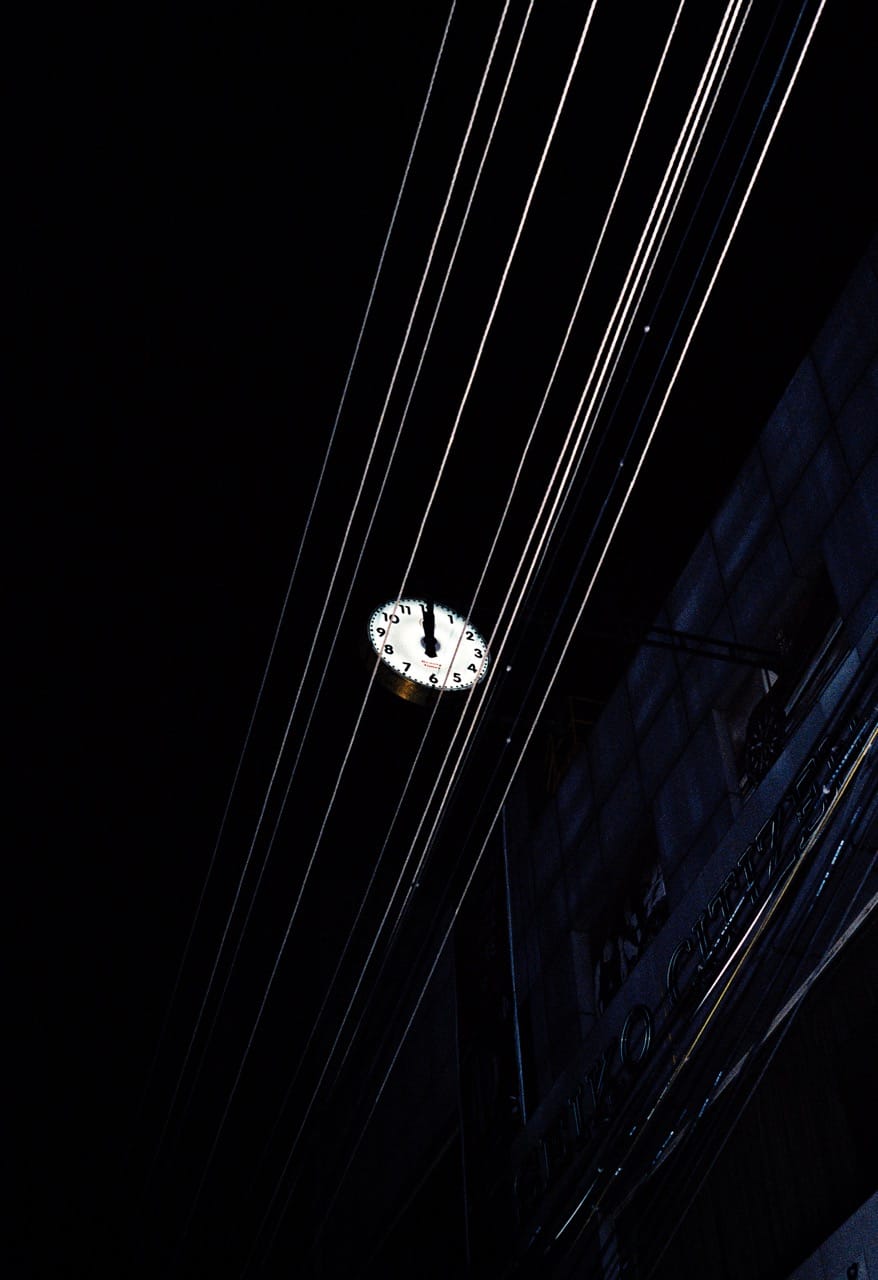 A clock illuminated at night is partially obscured by power lines, set against a dark building and sky.