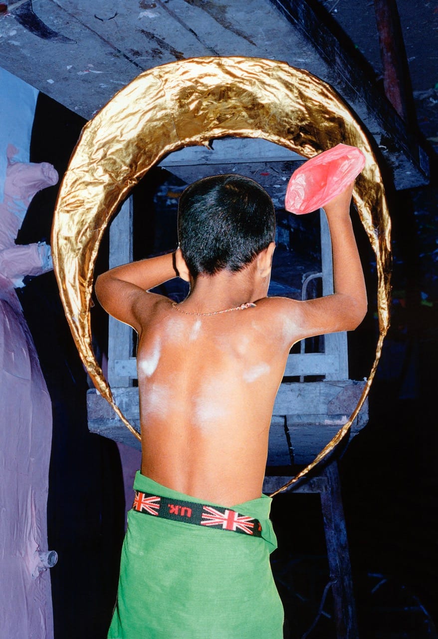 A child with a painted back holds a red object, standing inside a golden hoop, wearing a green garment with a Union Jack waistband.