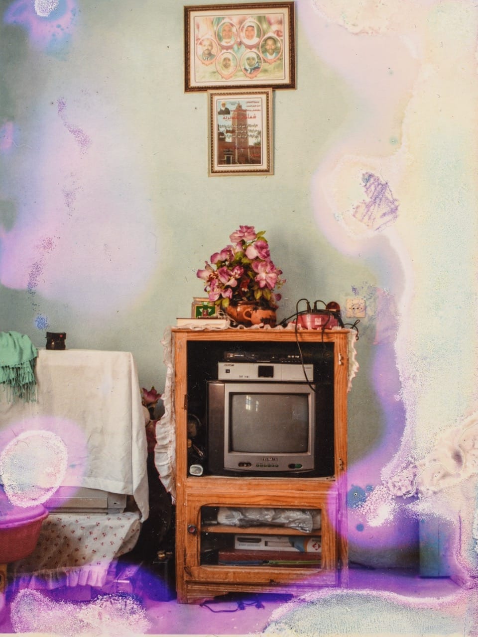 Vintage room with an old TV on a wooden stand, floral decor, and framed art on a pastel wall. The photo has colorful, abstract stains.