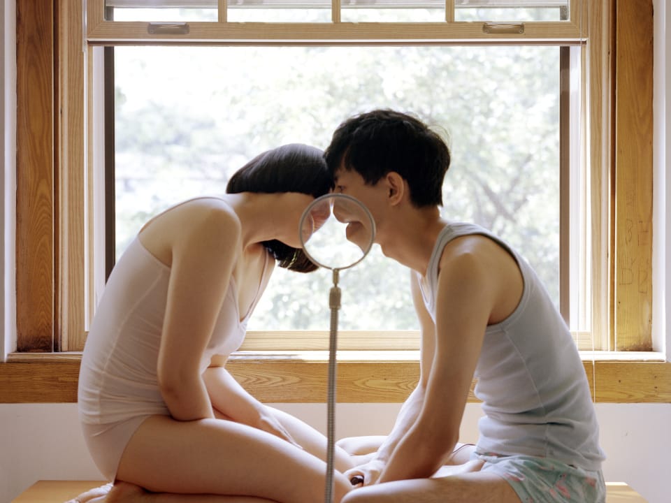 Two people sit on a windowsill facing each other, leaning in to touch lips through a standing magnifying glass.