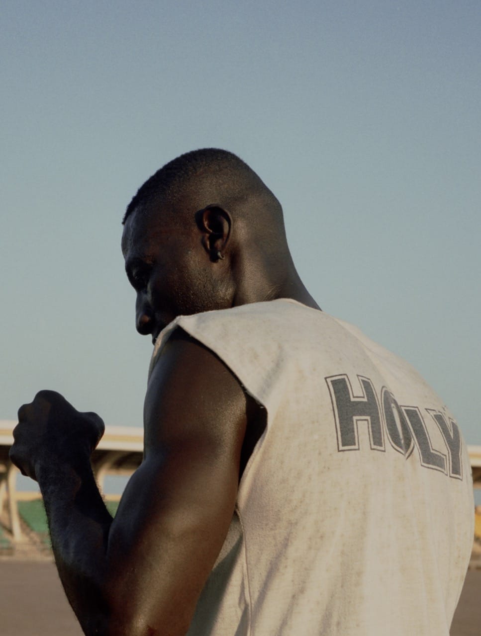 A person in a sleeveless shirt with "HOLY" on the back stands outdoors, with a clear sky and a bridge in the background.