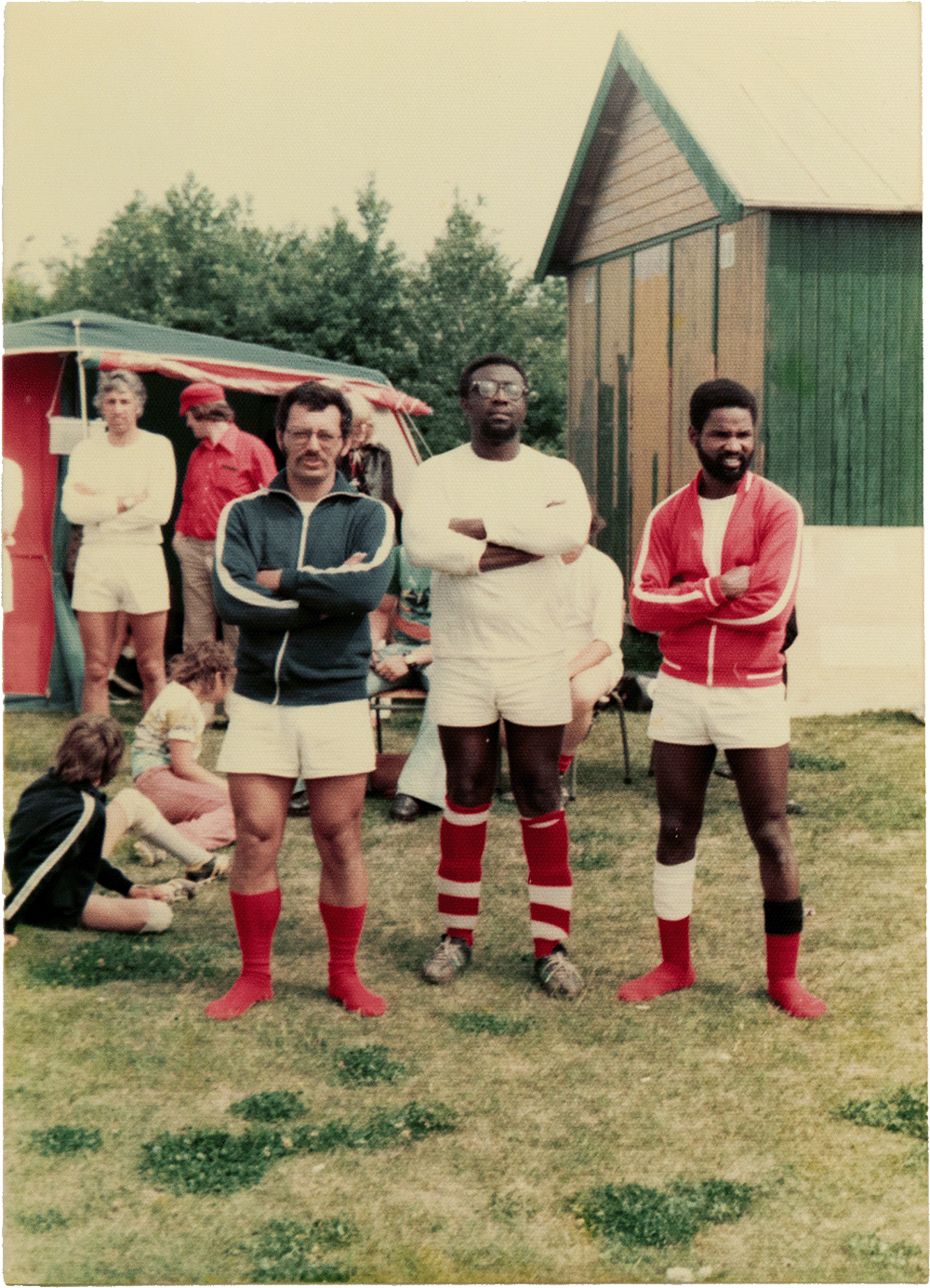 Archival photo showing three man (one white, two Black) in soccer outfits at a campground, with people in the background. © Sander Coers