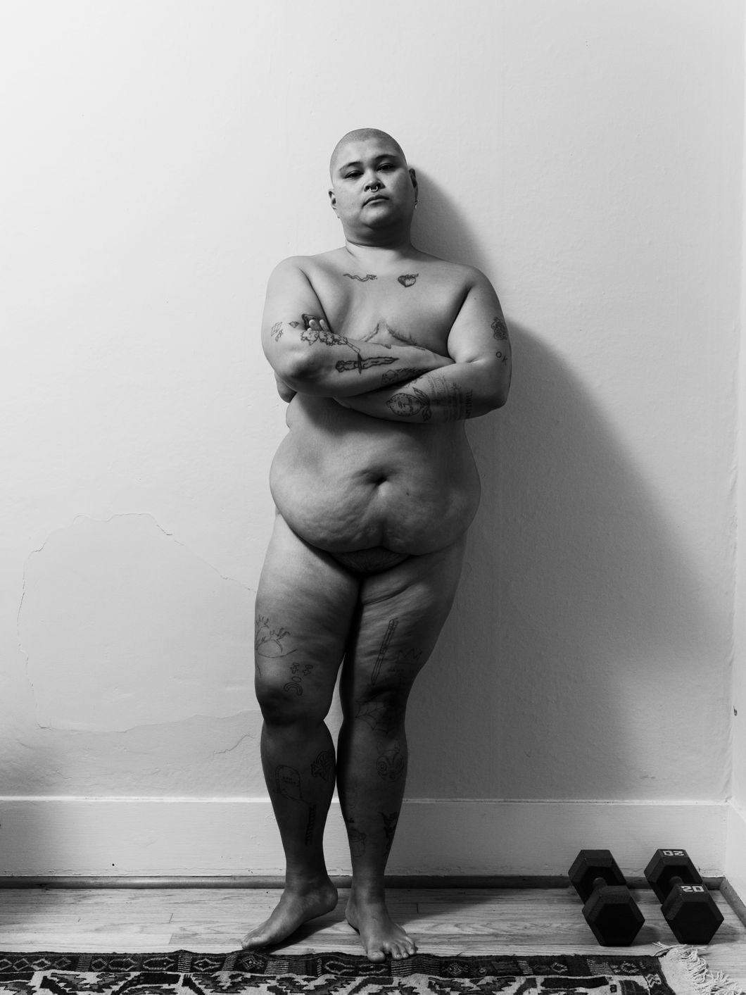 Black and white portrait of a nude person leaning against a wall. Two dumbbells lie on the floor besides them, next to a woven carpet. The person has a nose piercing, bald head and holds their tattooed arms crossed, looking strongly into the camera. © Ricardo Nagaoka