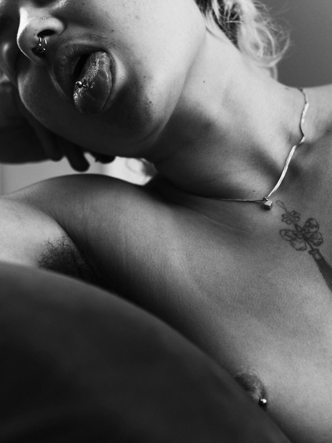 Black and white close-up of a topless person, showing half of their face, chest and throat. Their nipple, nose and tongue (sticking out) are pierced. They wear a thin necklace and their chest is tattooed. © Ricardo Nagaoka