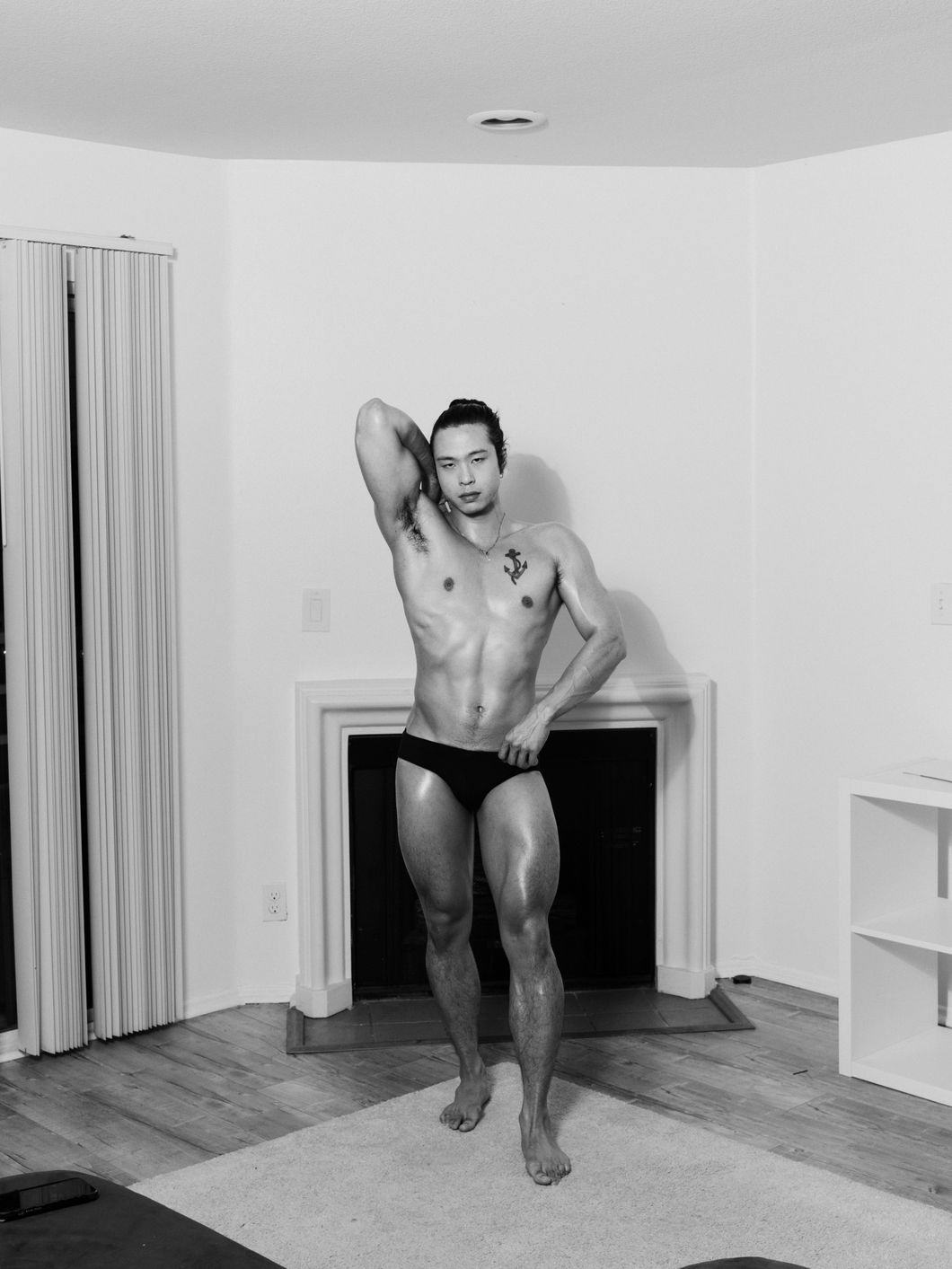 Black and white portrait of a man, with an Asian appearance, and trained physique. They pose with one arm stretched upwards and back, revealing the right armpit and tattooed chest. They stand in a living room in front of an empty fire place, on a rug © Ricardo Nagaoka