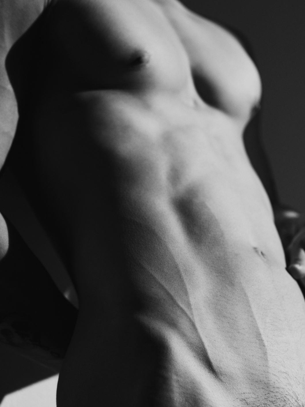 Black and white close-up of a nude male torso, showing chest and belly. The light comes in from the right, creating a shadow across the torso on the left. © Ricardo Nagaoka