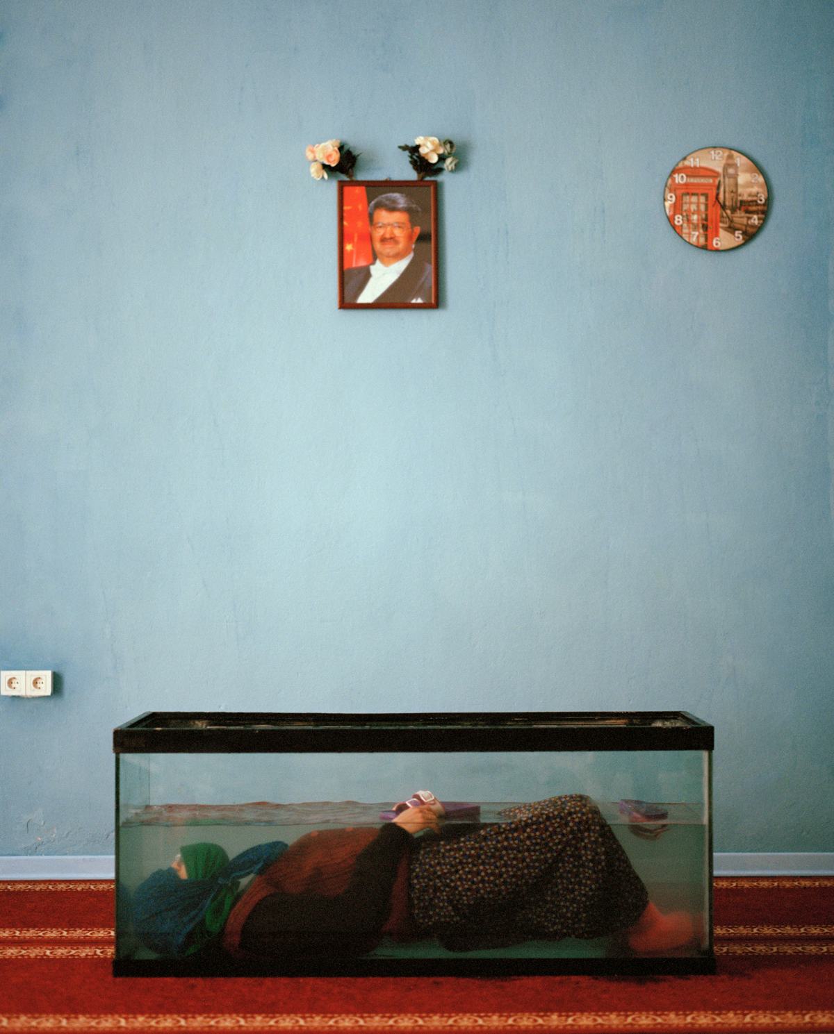 A women dressed in traditional Anatolian clothing lying in a fish tank, in a what appears to be a Turkish home (red carpet on the floor, blue walls.) On the wall hangs a portrait of Turgut Özal and a clock showing the Big Ben in London.