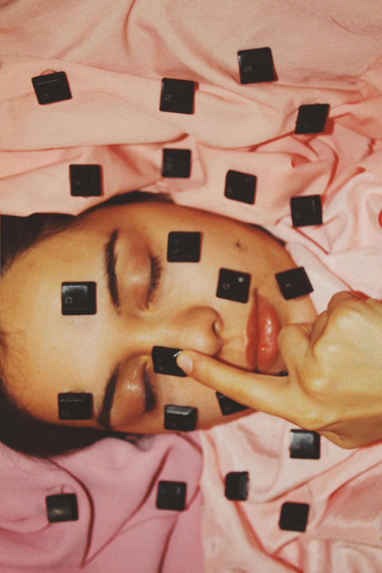 Portrait of a woman surrounded by pink fabric with computer keys placed over her face. © Alp Peker