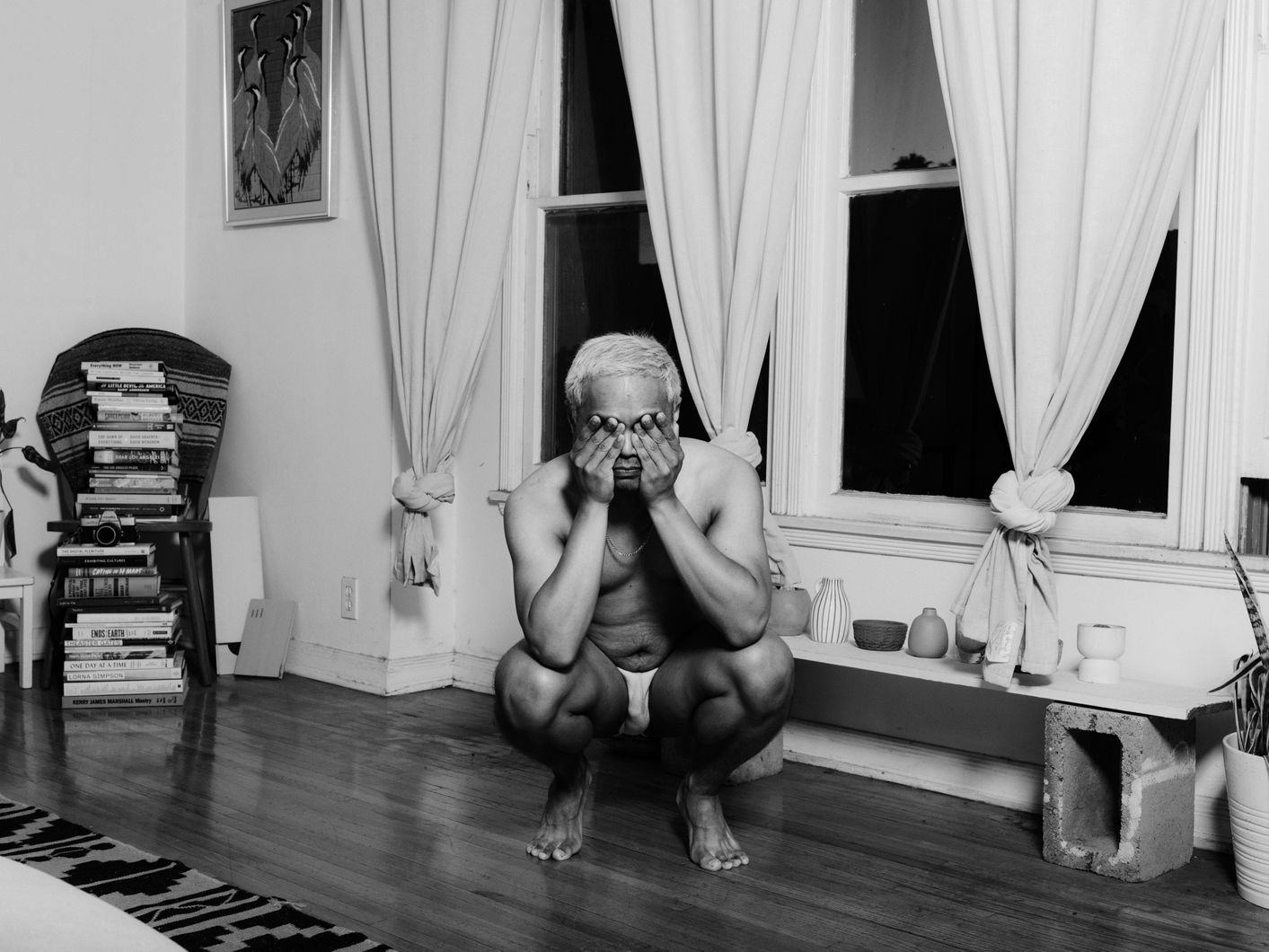 Black and white image of a person squatting in a living room. The person is wearing white underwear and a necklace, has bleached hair and covers their eyes with their hands. In the background, there are two windows with tied up curtains, a pile of books, some pottery and a painting. © Ricardo Nagaoka