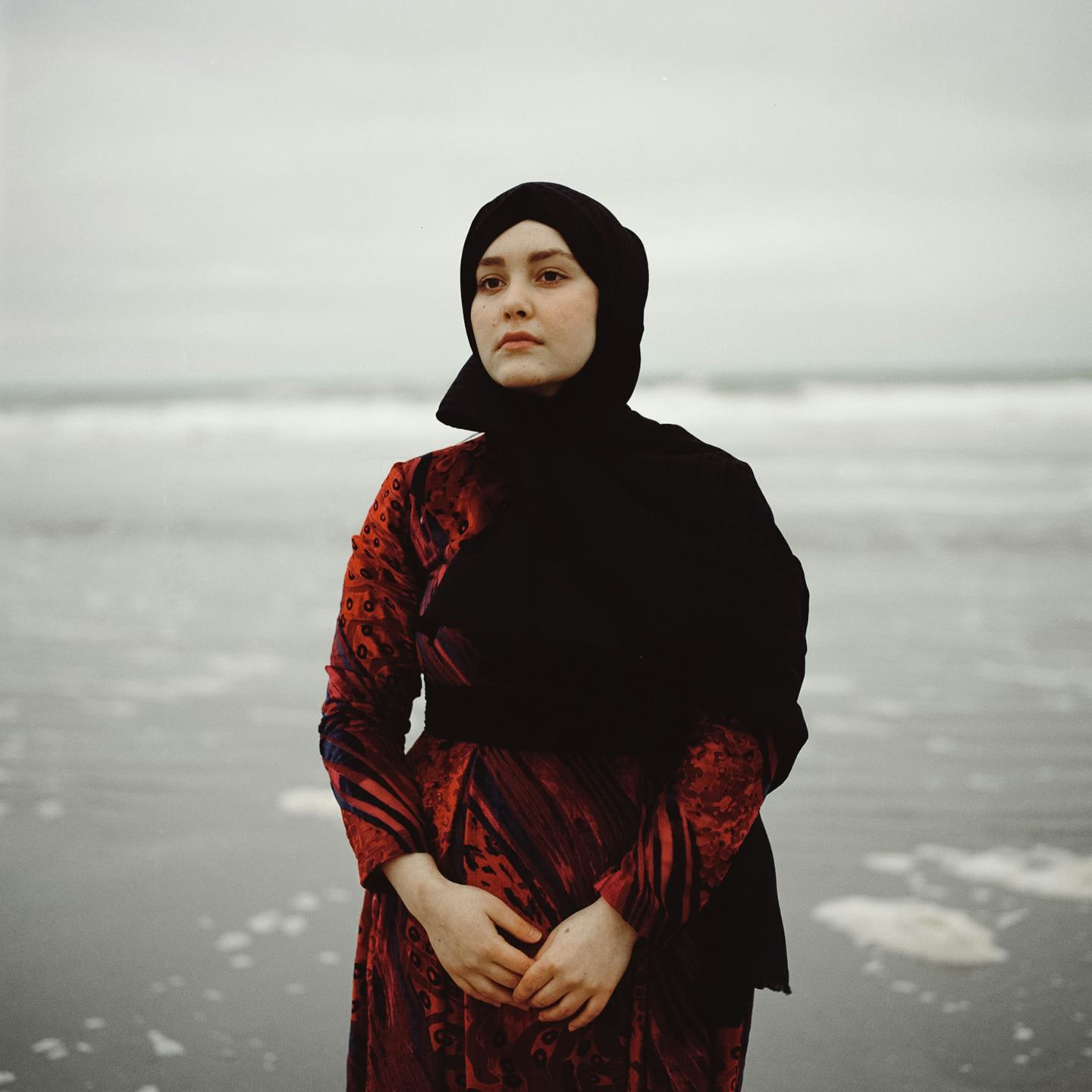Women wearing a red dress and a black hijab overlooks a grey Dutch sea