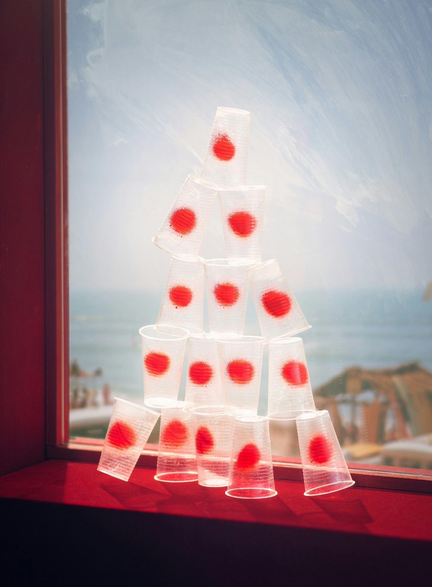 Image of a red window overlooking the ocean. On the window sill is a pyramid of stacked plastic cups, each with a large red dot.