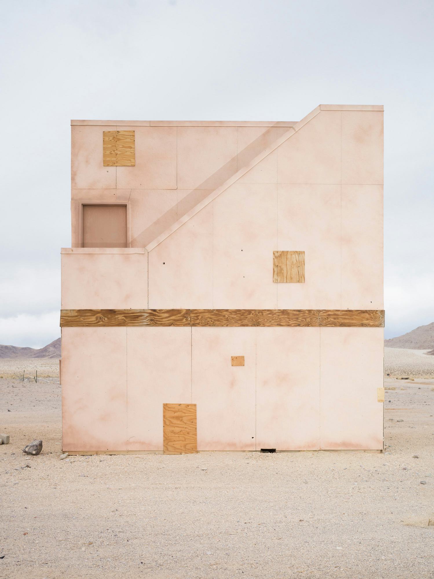 Image of a beige-coloured concrete facade of a building in a desert, with boarded windows © Andrea Orejarena & Caleb Stein