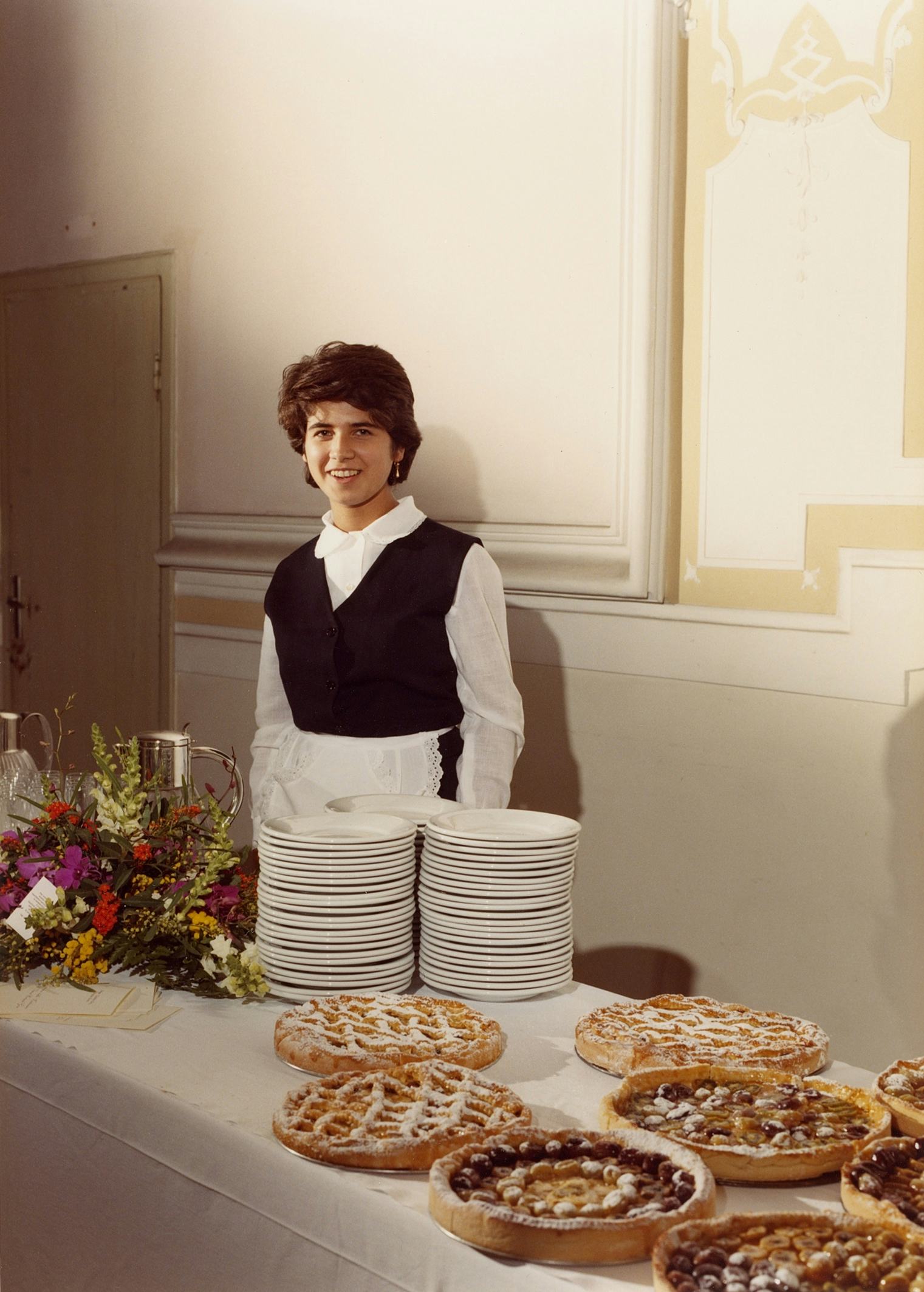Archival image of the artist's mother, as a waitress, standing behind a table full of flowers, plates and pies. © Eleonora Agostini