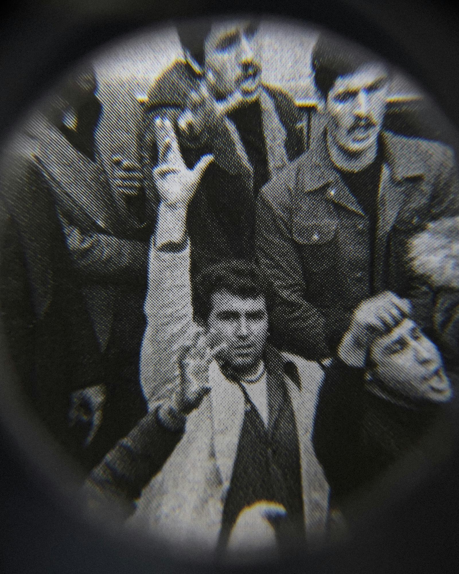 Black and white close-up image of an existing photograph, showing a man in the middle of a protesting crowd staring into the camera, raising his arm and hand. Shot through a magnifying lens © Amin Yousefi