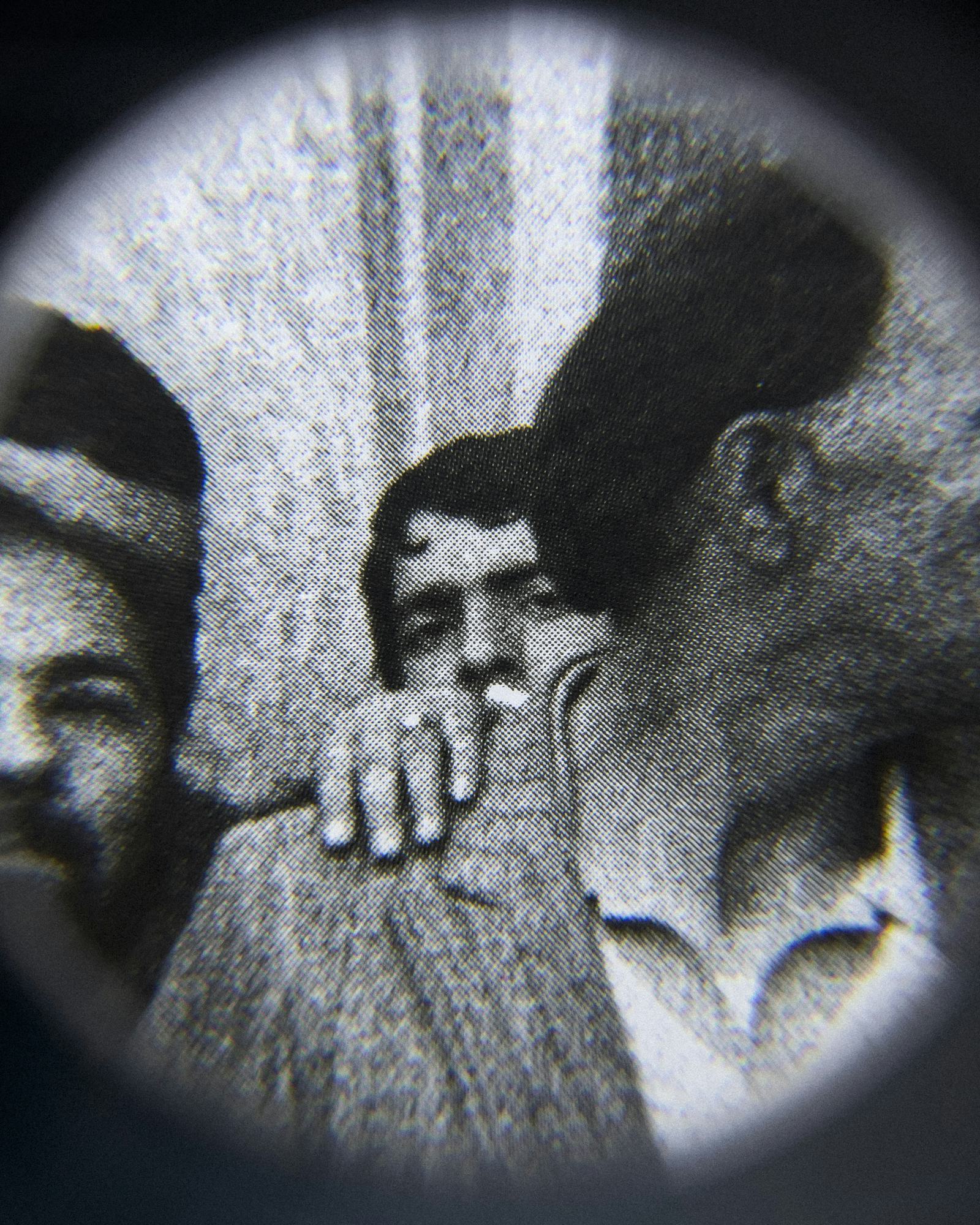 Black and white close-up image of an existing photograph, showing a man peeking into the camera from behind another man. Shot through a magnifying lens © Amin Yousefi
