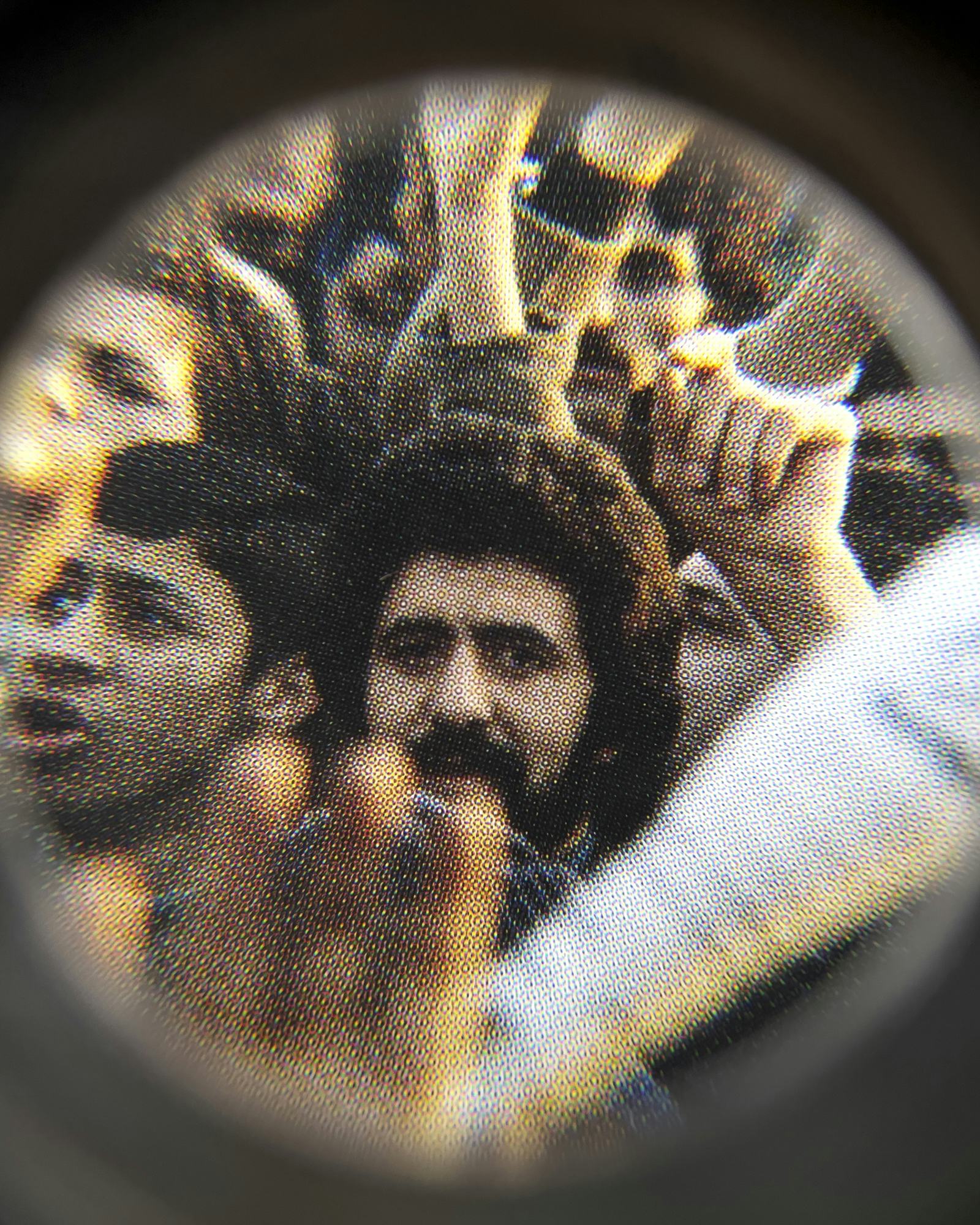Coloured close-up image of an existing photograph, showing a man in the middle of a protesting crowd staring into the camera. Shot through a magnifying lens © Amin Yousefi