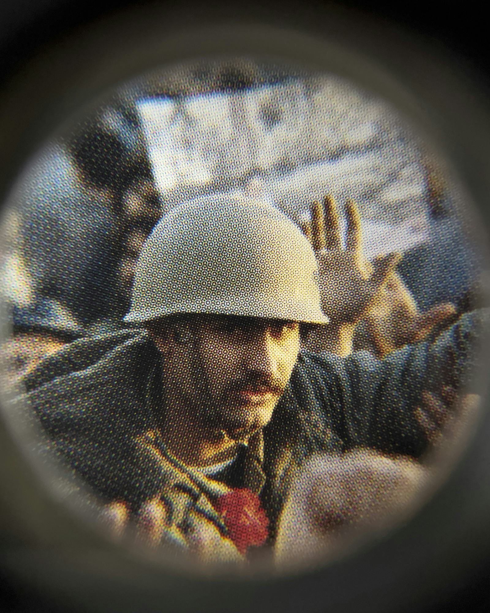 Coloured close-up image of an existing photograph, showing a man wearing a helmet staring into the camera, in the middle of a protest. Shot through a magnifying lens © Amin Yousefi