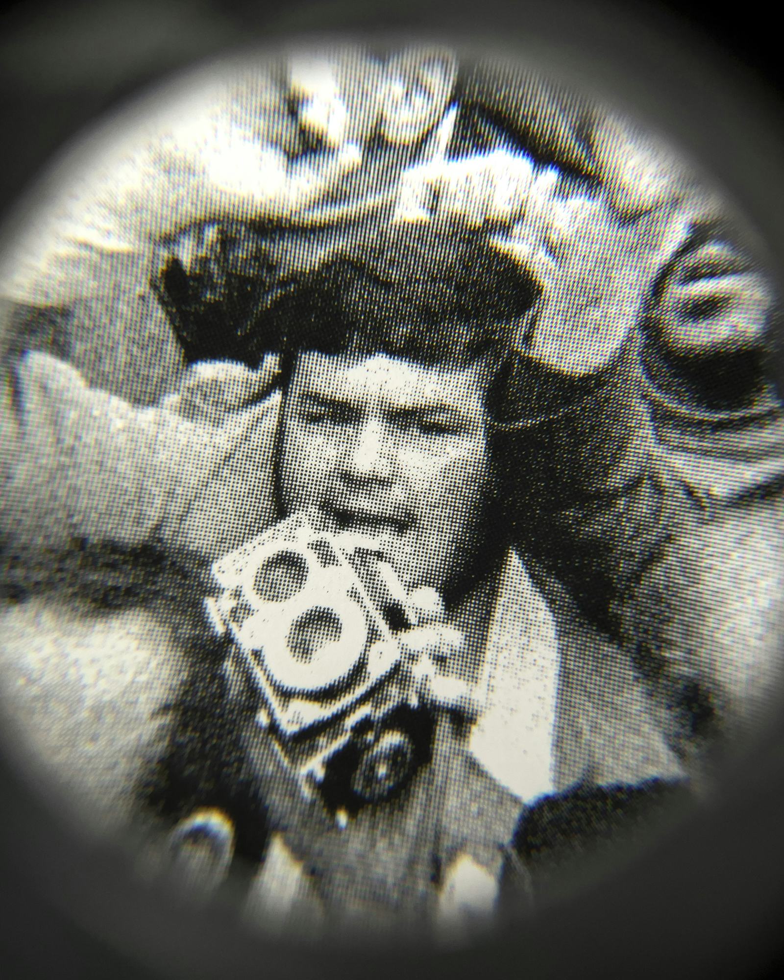 Black and white close-up image of an existing photograph, showing a man, holding a camera in the middle of a protesting crowd, staring into the camera. Shot through a magnifying lens © Amin Yousefi