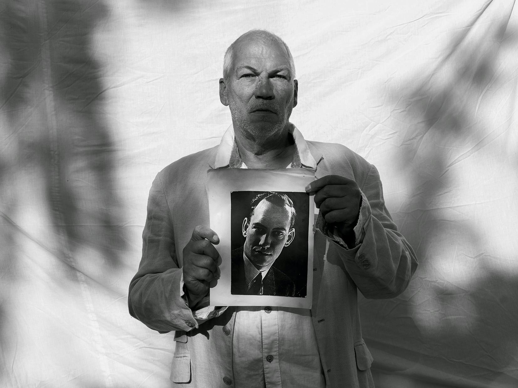 Black and white portrait of a white man, standing in front of a white sheet on which shadows are cast from surrounding trees. The man holds a historic portrait of another man. © Bebe Blanco Agterberg