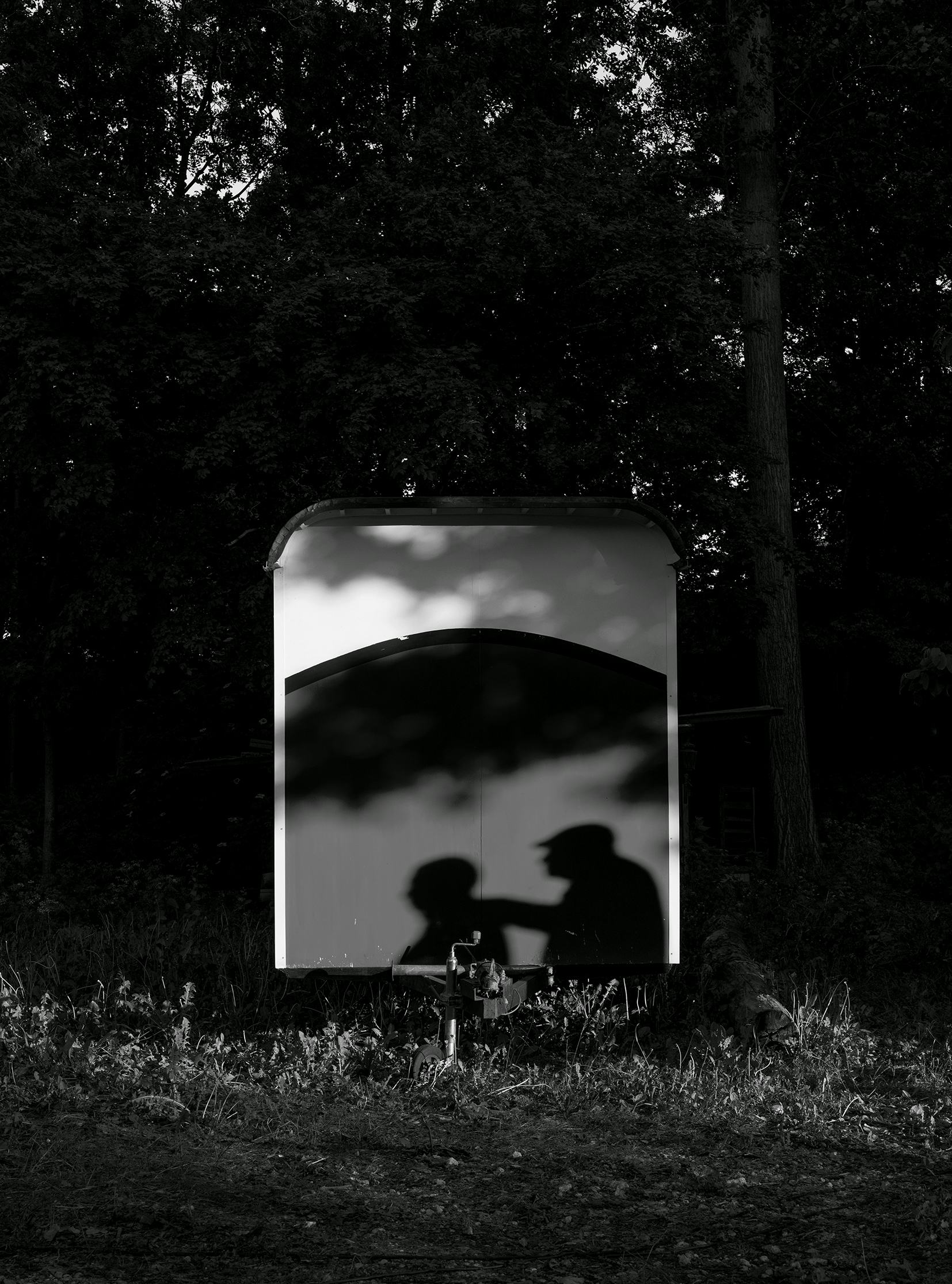 Black and white image of a trailer in a forest environment, on which the shadow of two men is visible. © Bebe Blanco Agterberg