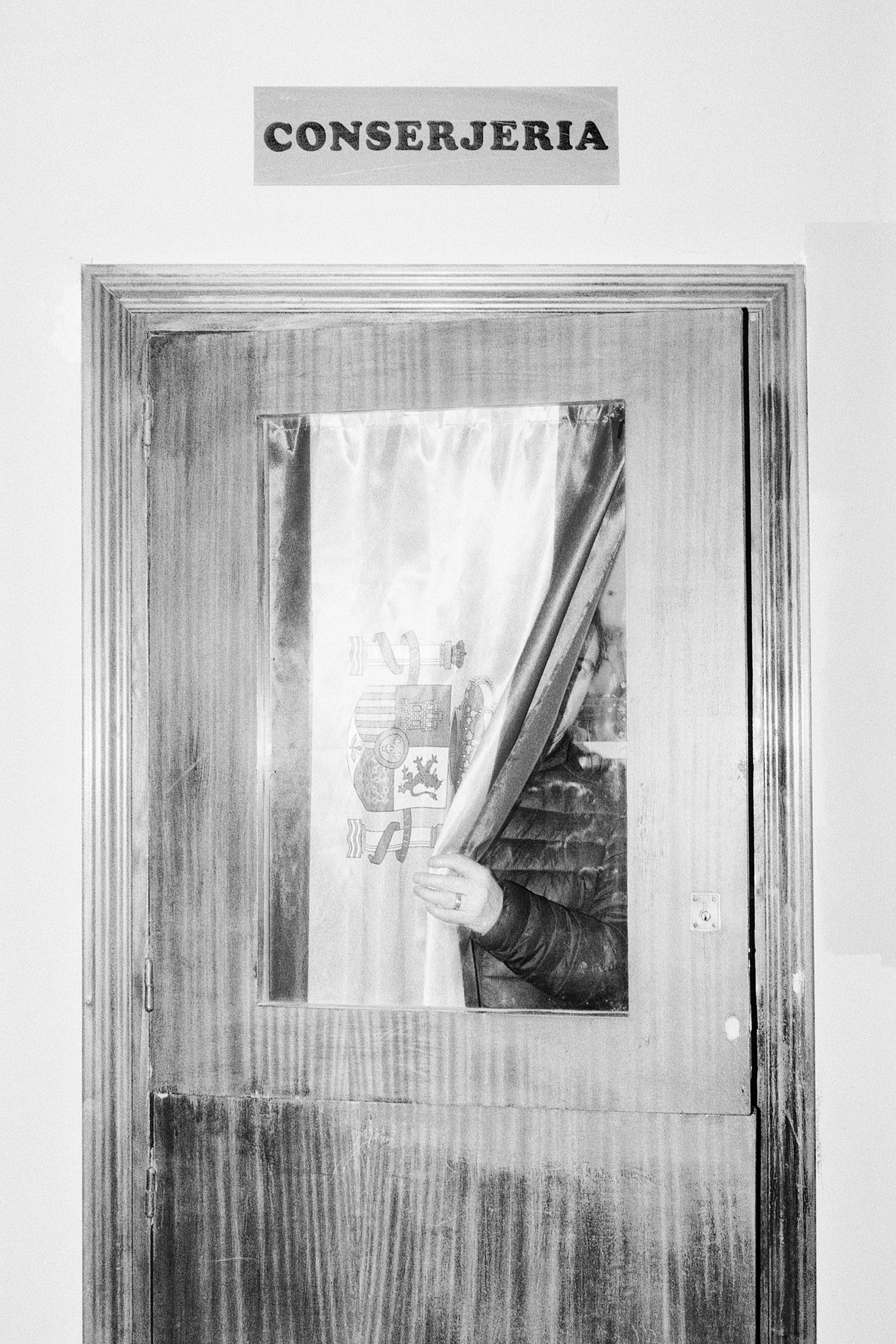 Black and white photo of a wooden door with a window covered by a Spanish flag from the Franco dictatorship. A person is partially visible behind the curtain. © Ignacio Navas