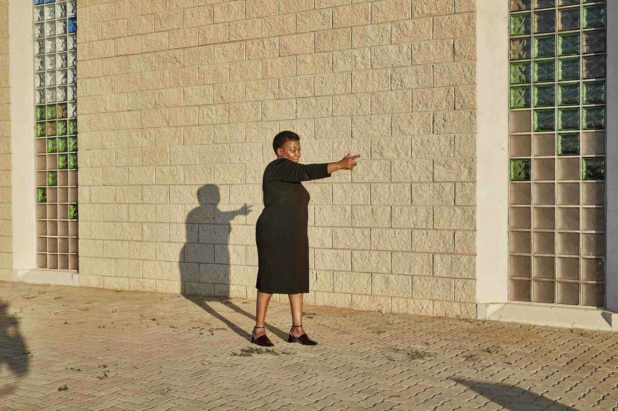 Photograph of a Black woman in front of a stone wall, pointing her finger as if it were a gun © Thero Makepe