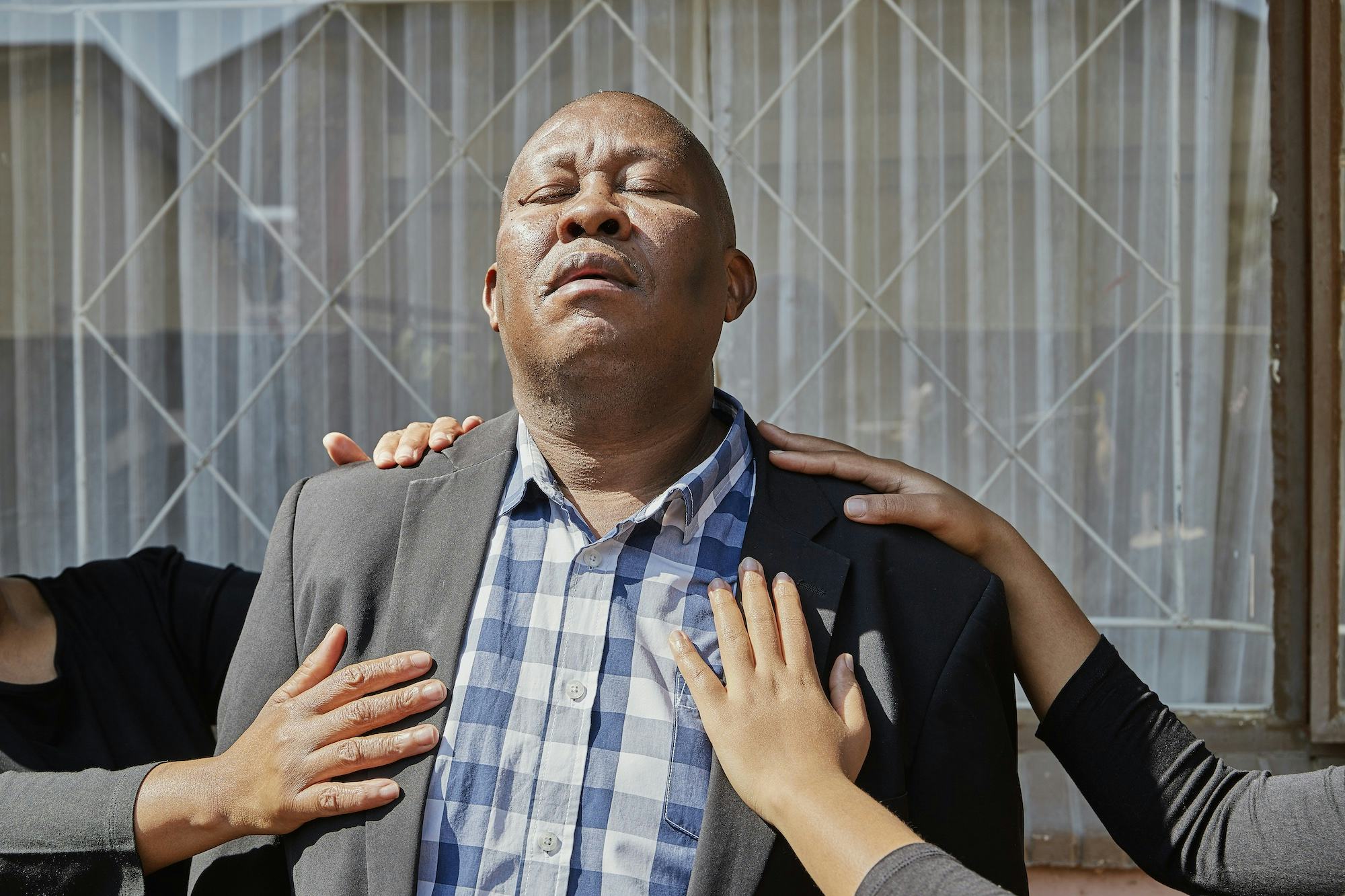 Portrait of a Black man in a suit, with his eyes closed and his face turned up towards the sun. Different pairs of hands hold his chest © Thero Makepe