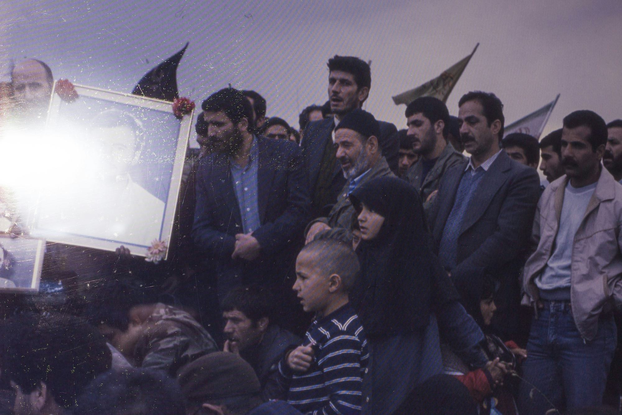 Photograph of a group of Iranian men and children, holding flags and the portrait of a martyr, obscured by a flash of light. © Ghazaleh Rezaei