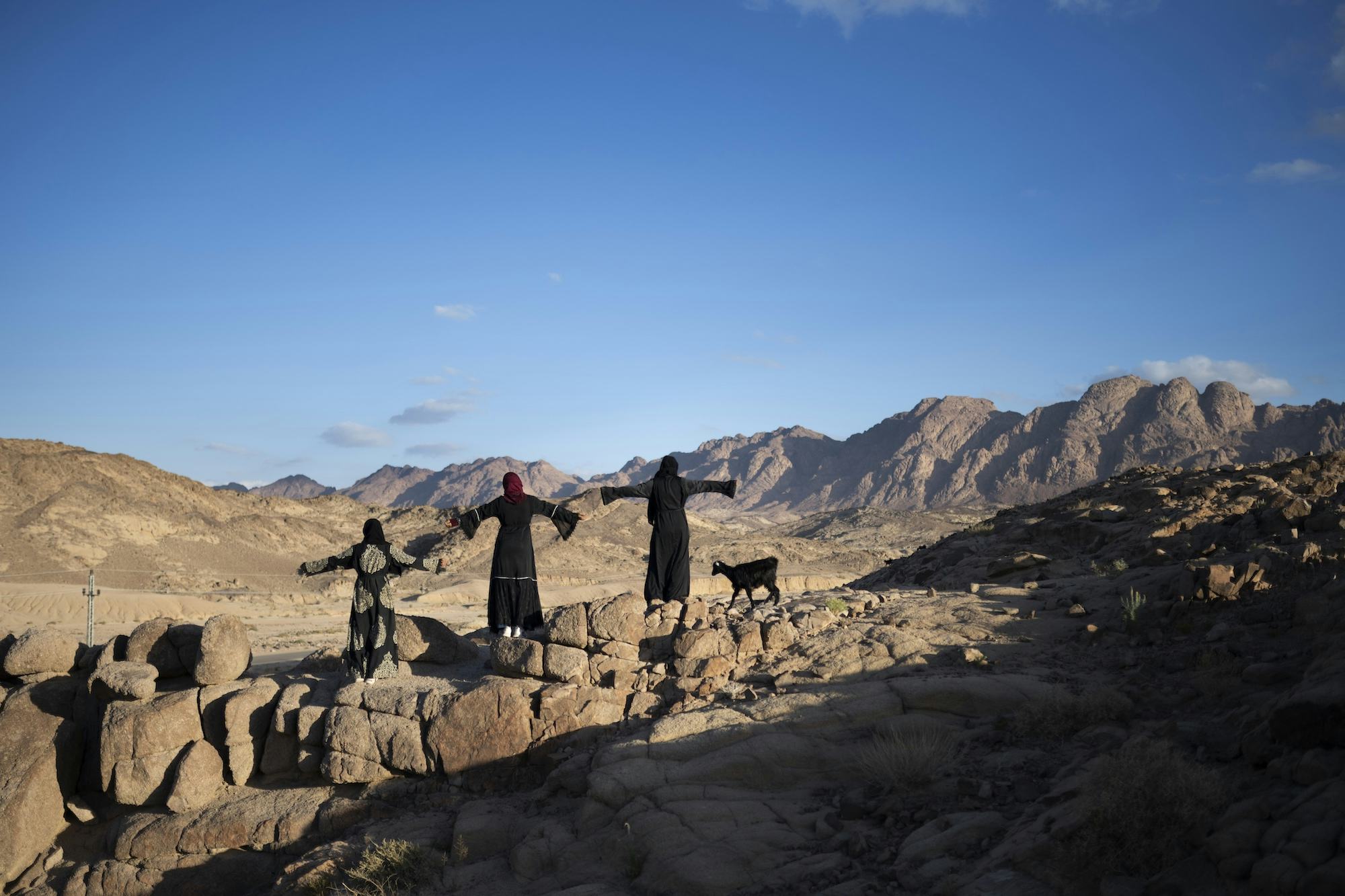 Three women of the Bedouin community in South Sinai, Egypt, herd their sheep across the moutains. They stand in a line with their arms spread out, overseeing the valley. © Rehab Eldalil