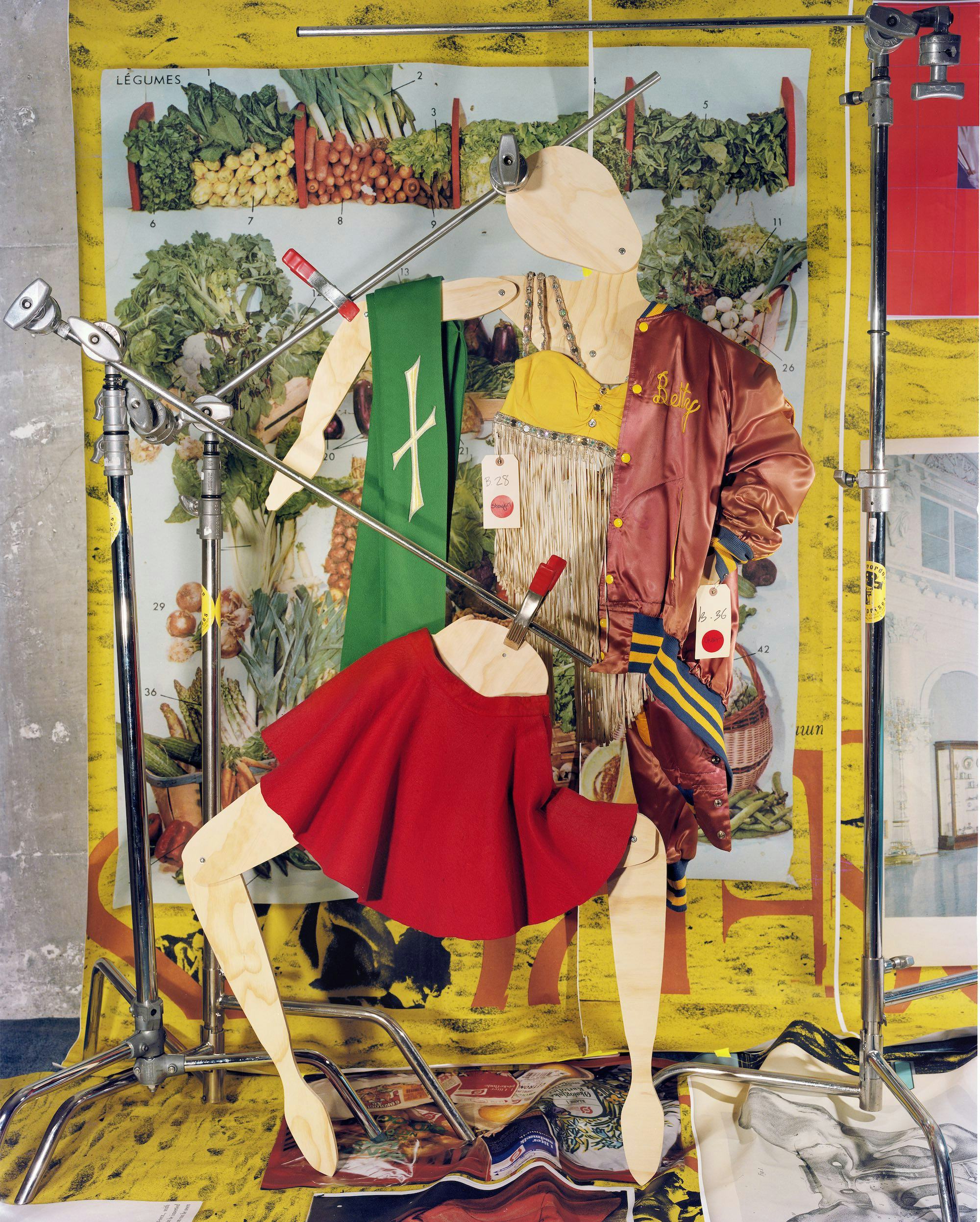 Photo of an abstract wooden mannequin dressed as a cheerleader, in front of a wallpaper showing vegetables.