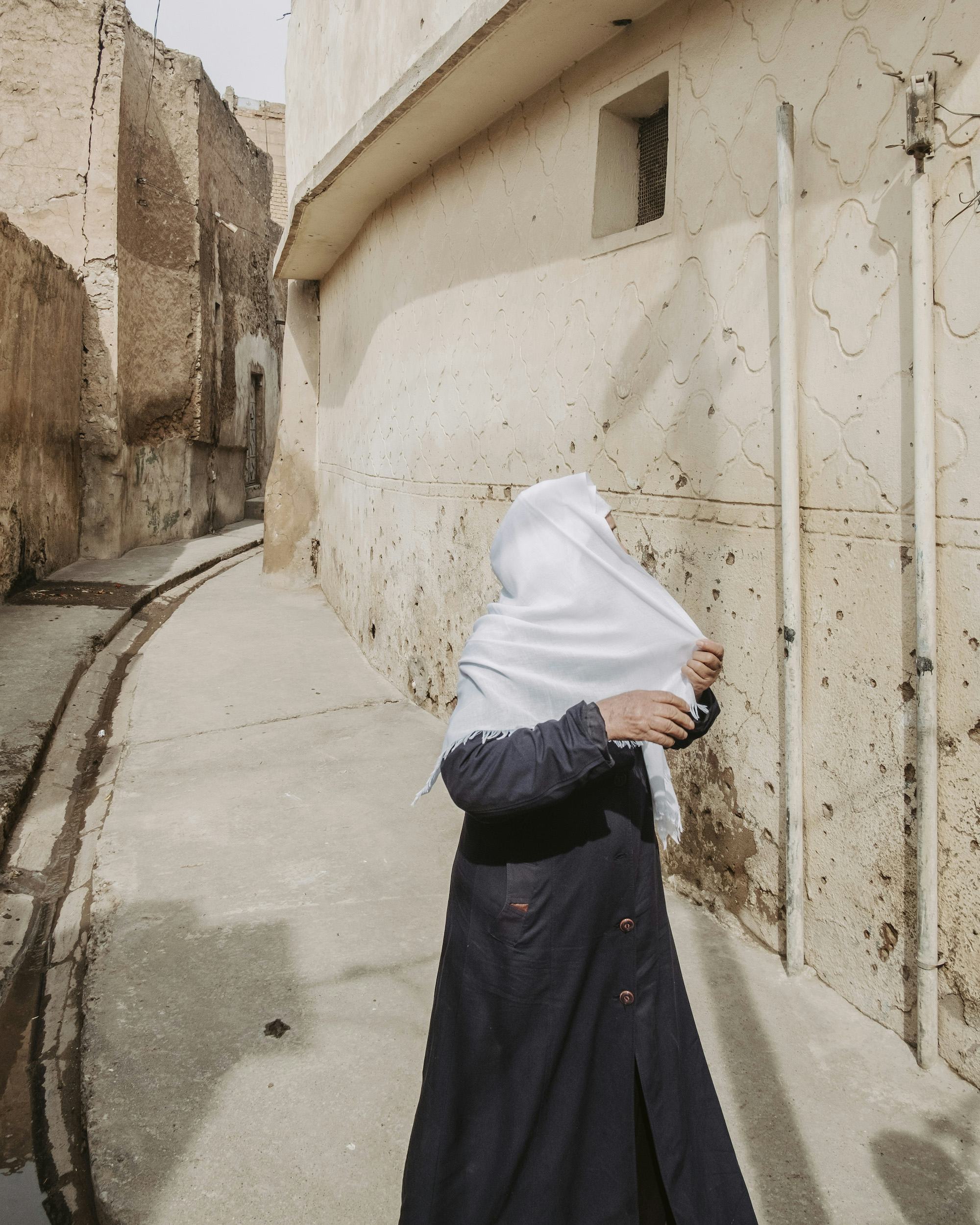 Image of an Iraqi woman in a black robe on an empty street, wearing a white hijab. Her face is turned away and hidden behind the hijab she pulls over her face.