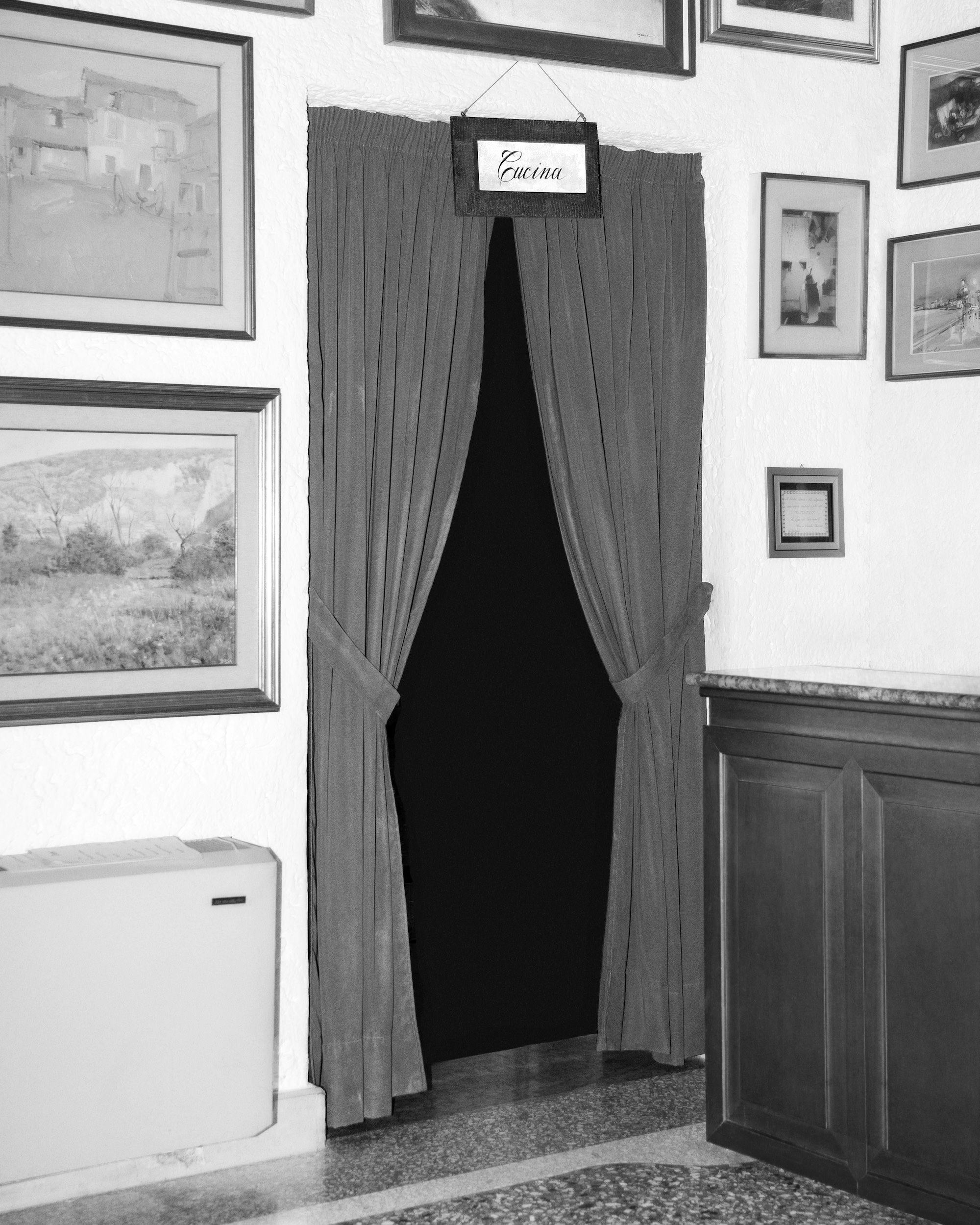 Black and white image of a doorway, framed by curtains, marked by a sign reading 'Cucina' The walls around are framed with various paintings and photographs in frames. © Eleonora Agostini