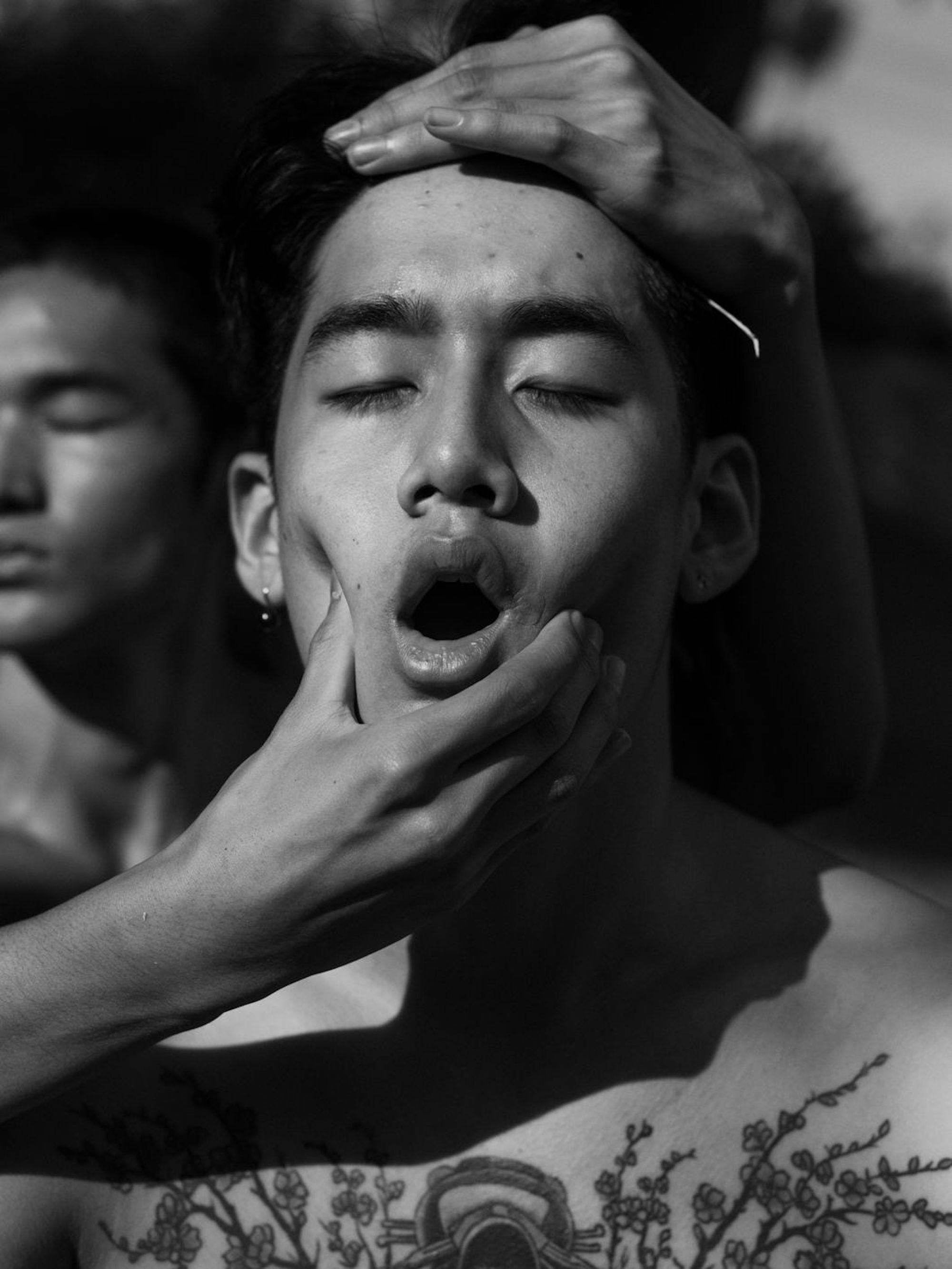 Black and white portrait of a man's face, eyes closed and mouth gaping. Part of his tattooed chest is visible and he wears a silver earring in his right ear lobe. His face is framed by two hands, grasping his head and squeezing his cheeks. In the background, half of the face of another person is visible. © Ricardo Nagaoka