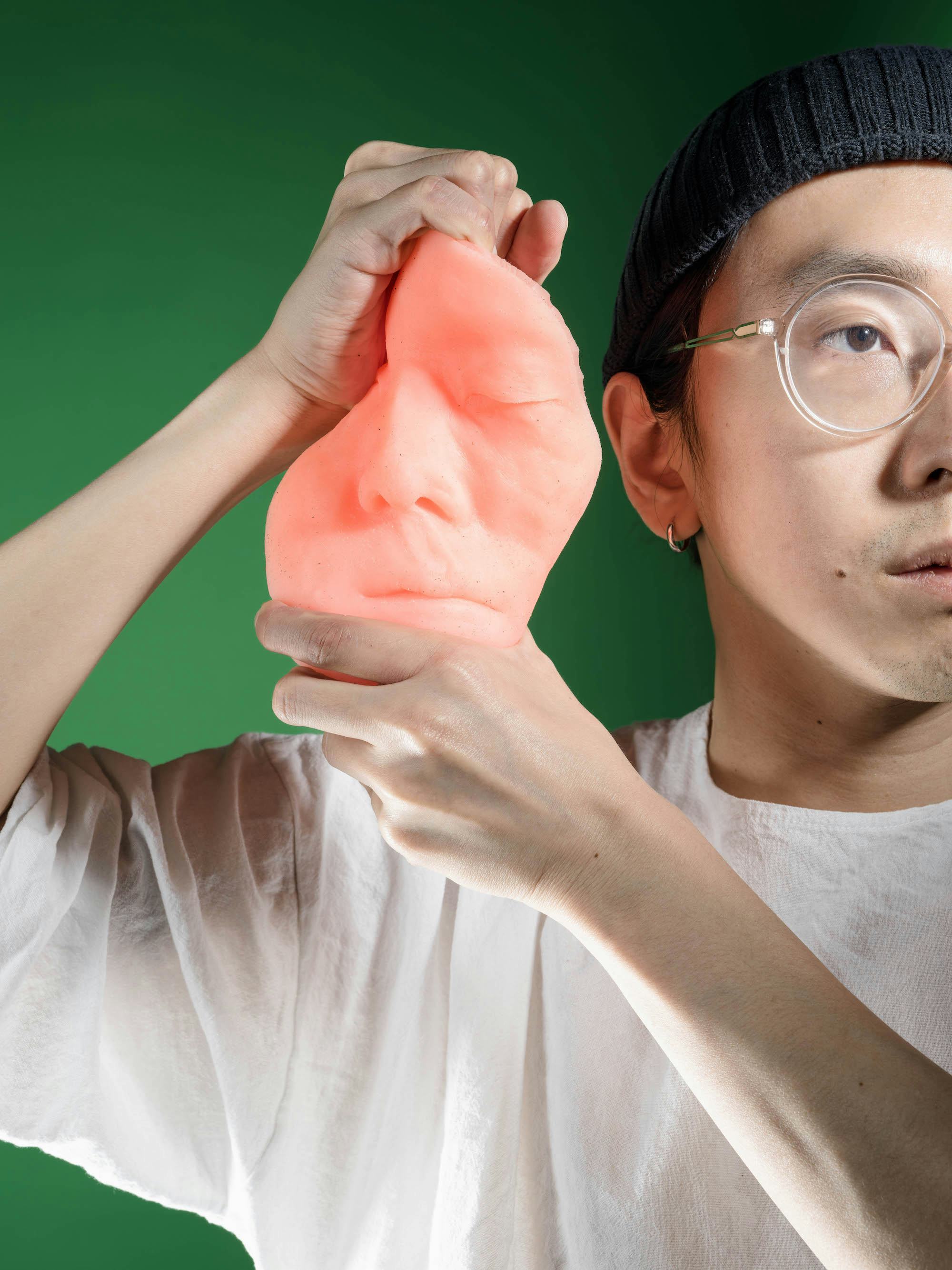 Pink silicon cast of the artist's face, in front of a green background, being twisted by the artist, holding it next to his face © Sheung Yiu