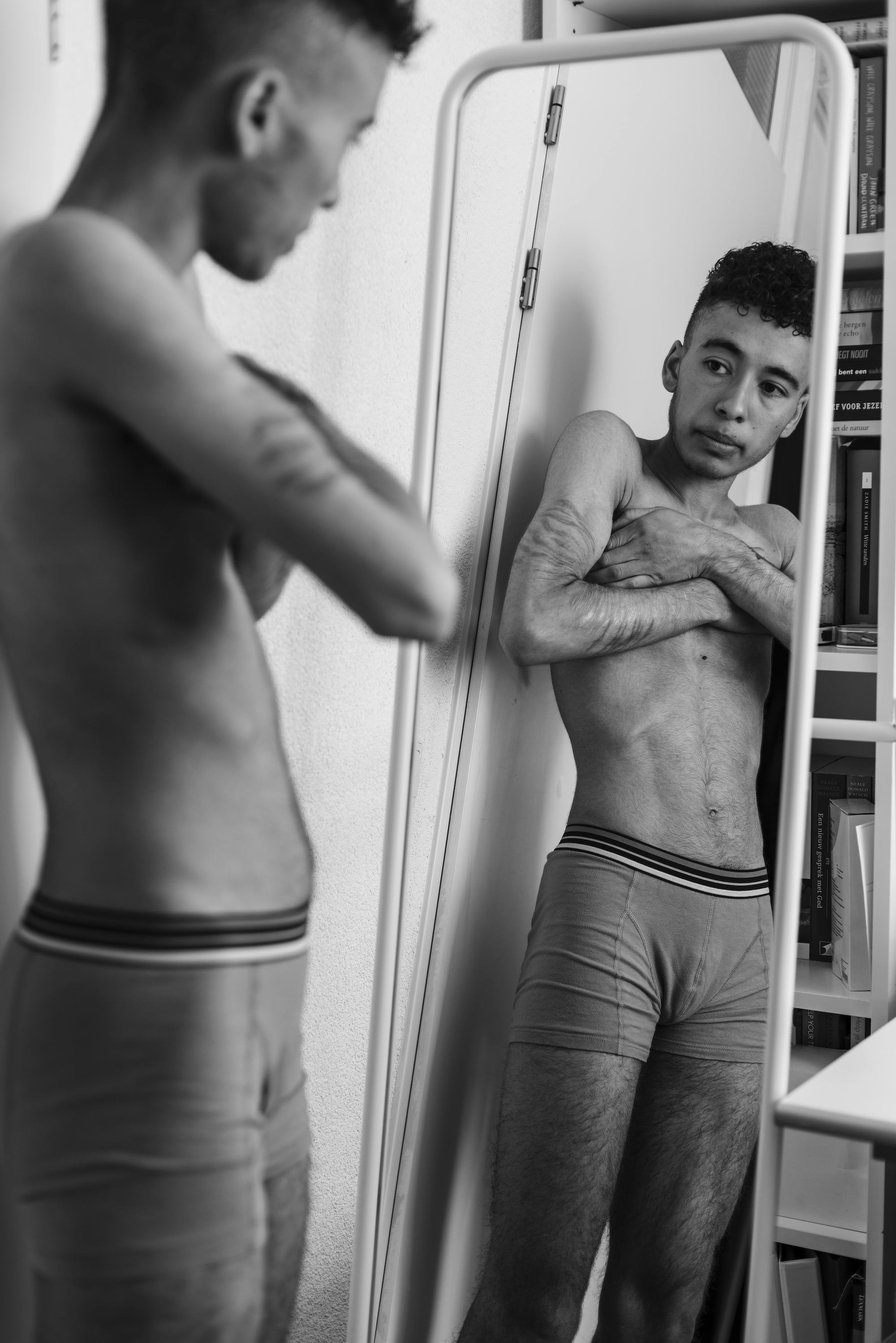 A black-and-white photo of the photographer, Marvel Harris, who stands topless in front of the mirror, clutching his arms around his chest.