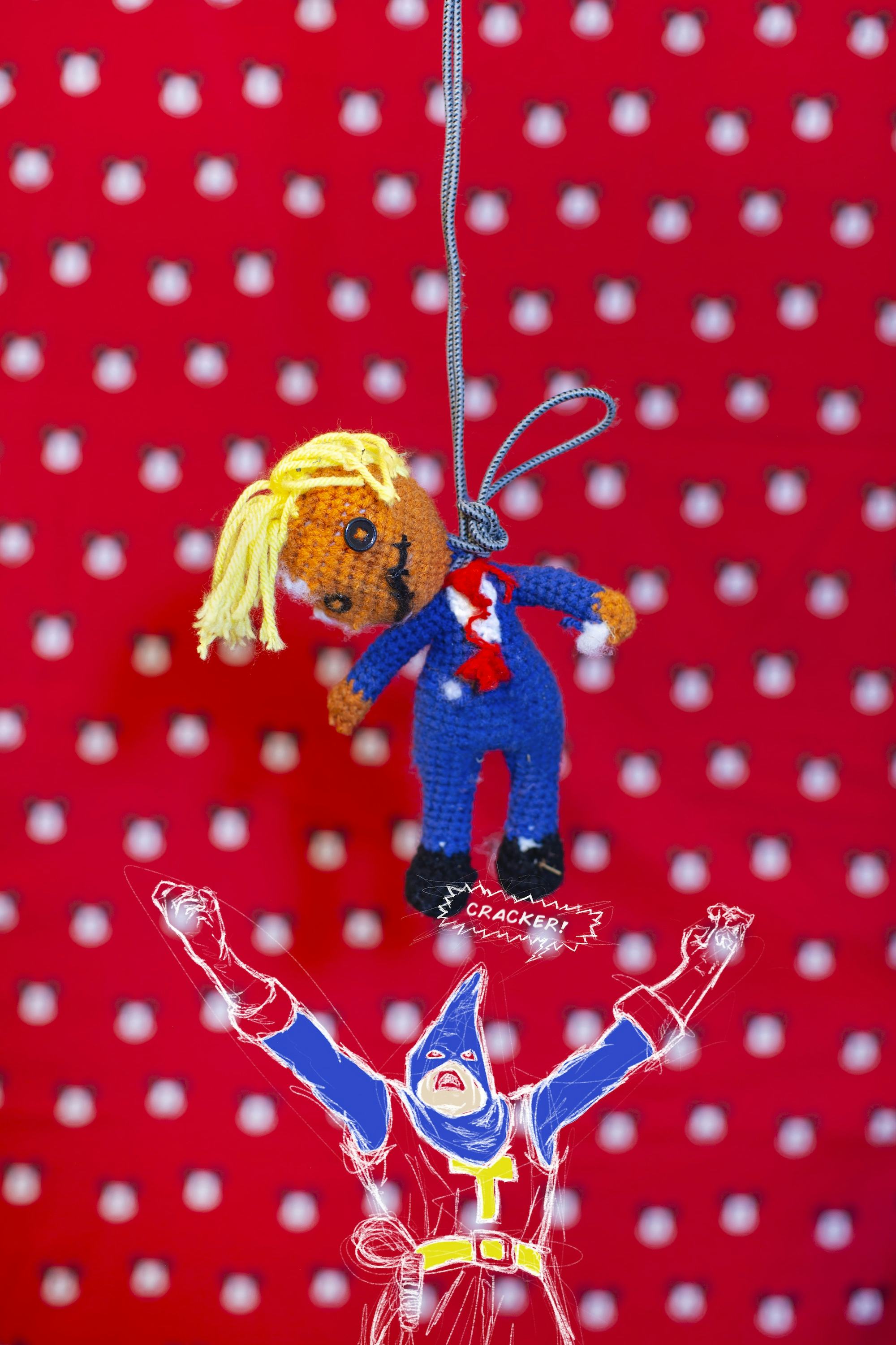 Image of a knitted puppet hanging by its neck on a rope in front of a red dotted background. © André Ramos-Woodard