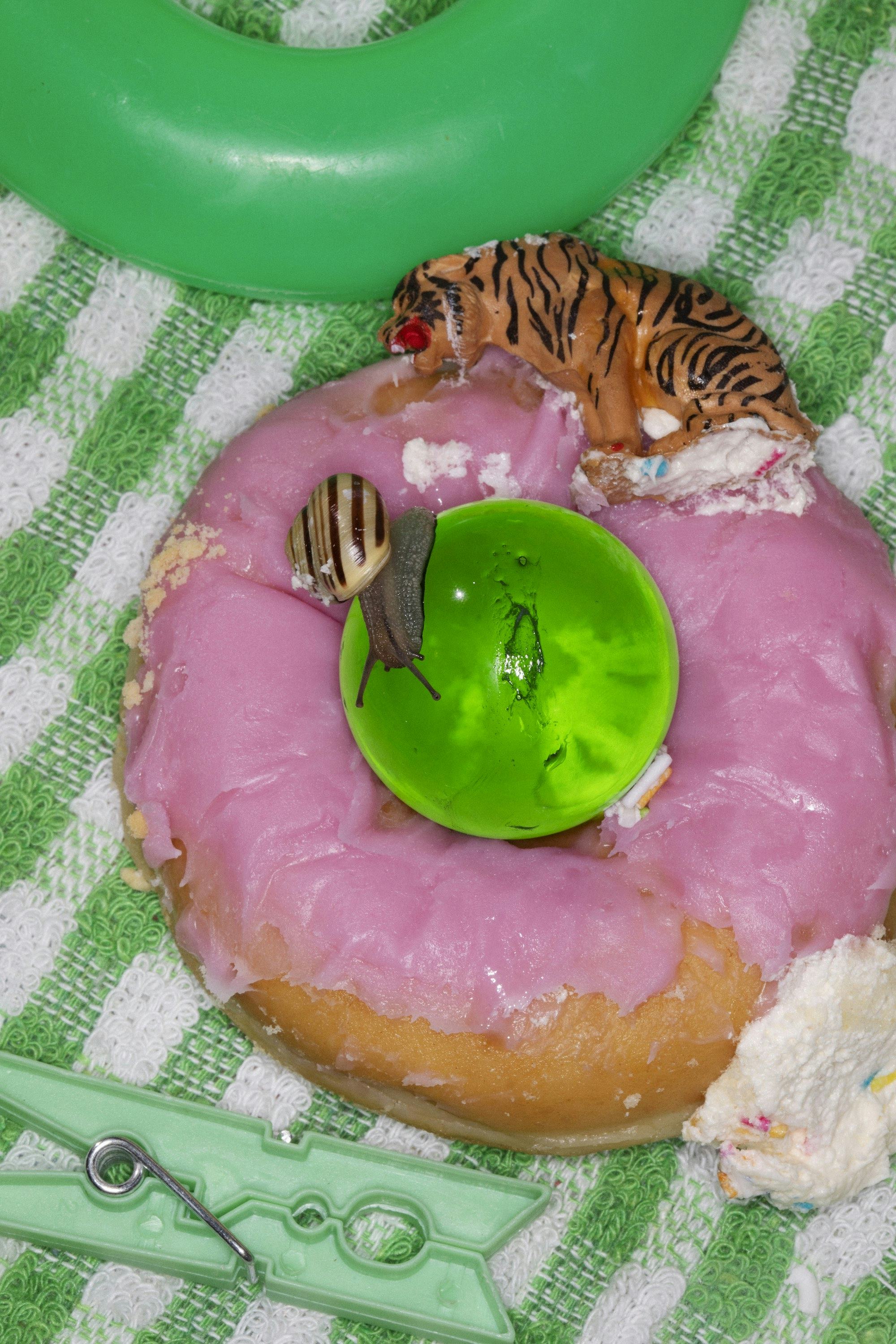 A photograph of a pink-glaced donut, on a green kitchen cloth, surrounded by a green cloth peg, a small toy tiger and a snail.
