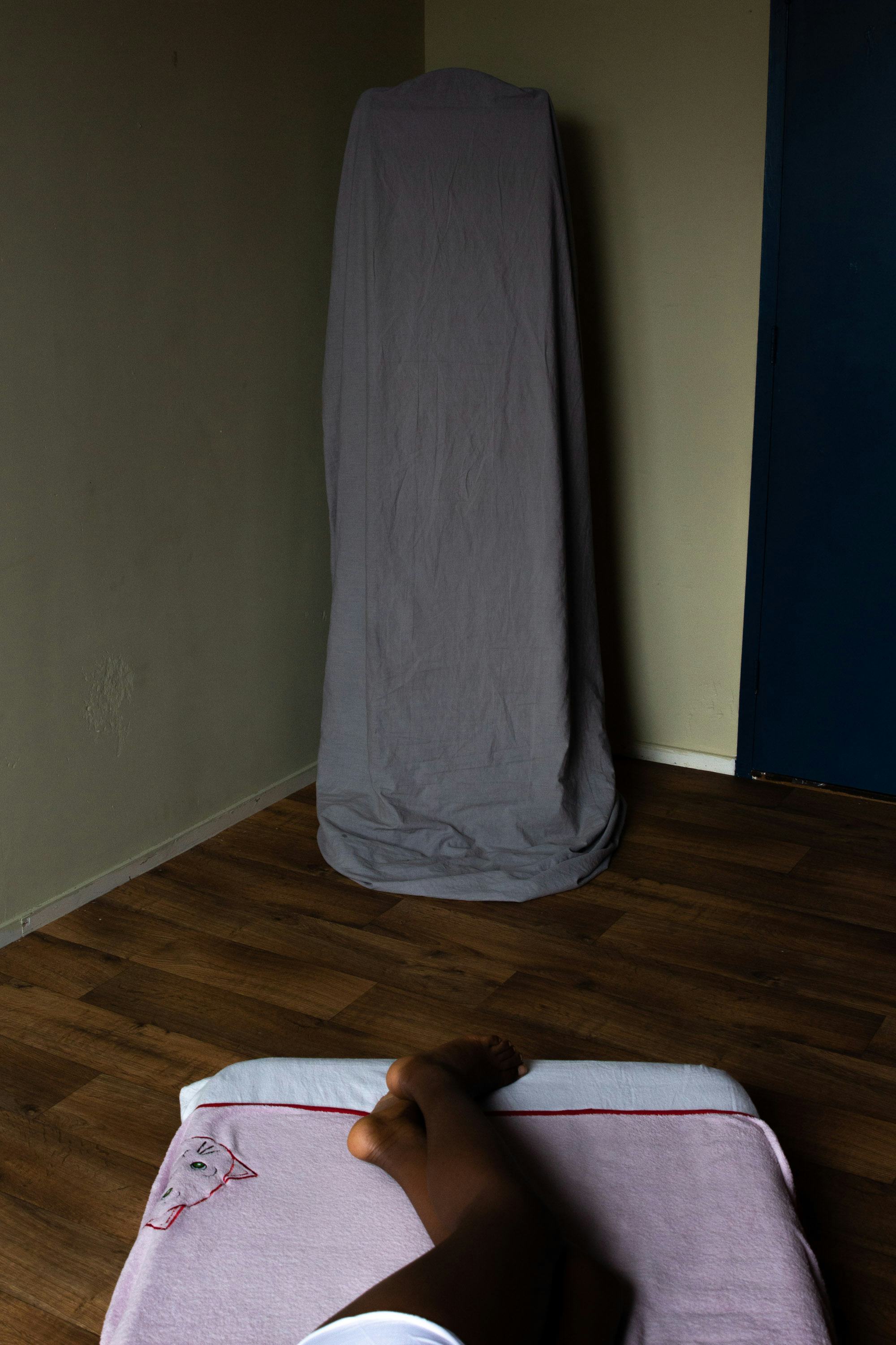 Photo of a black person's legs, lying on a mattress, with an object in the corner of the room covered by a sheet. © Iyanla Etnel